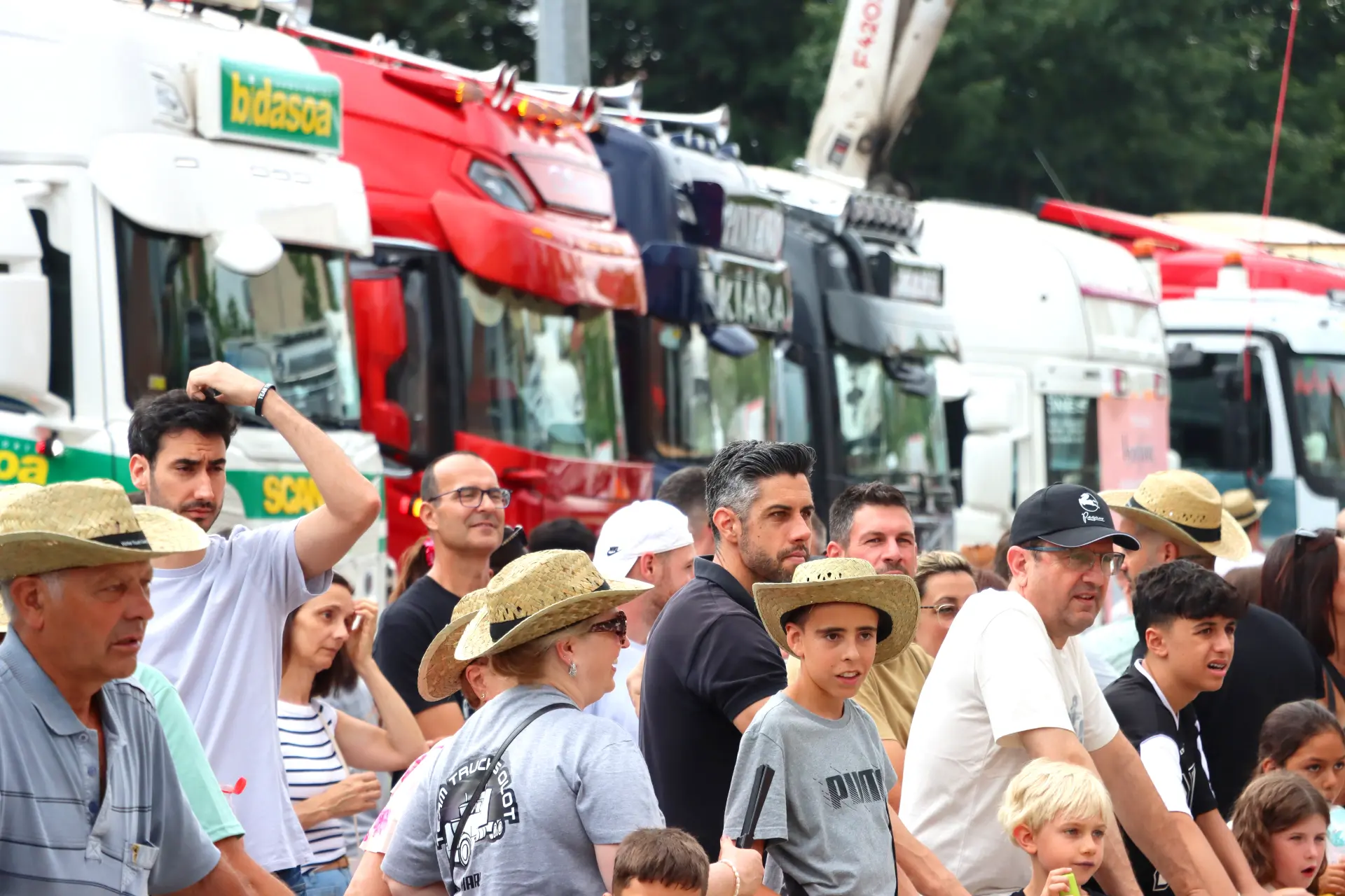 Una multitud de personas, algunas con sombrero, permanecen de pie frente a coloridos camiones aparcados en la XI Concentración de Camiones de Olot al aire libre.