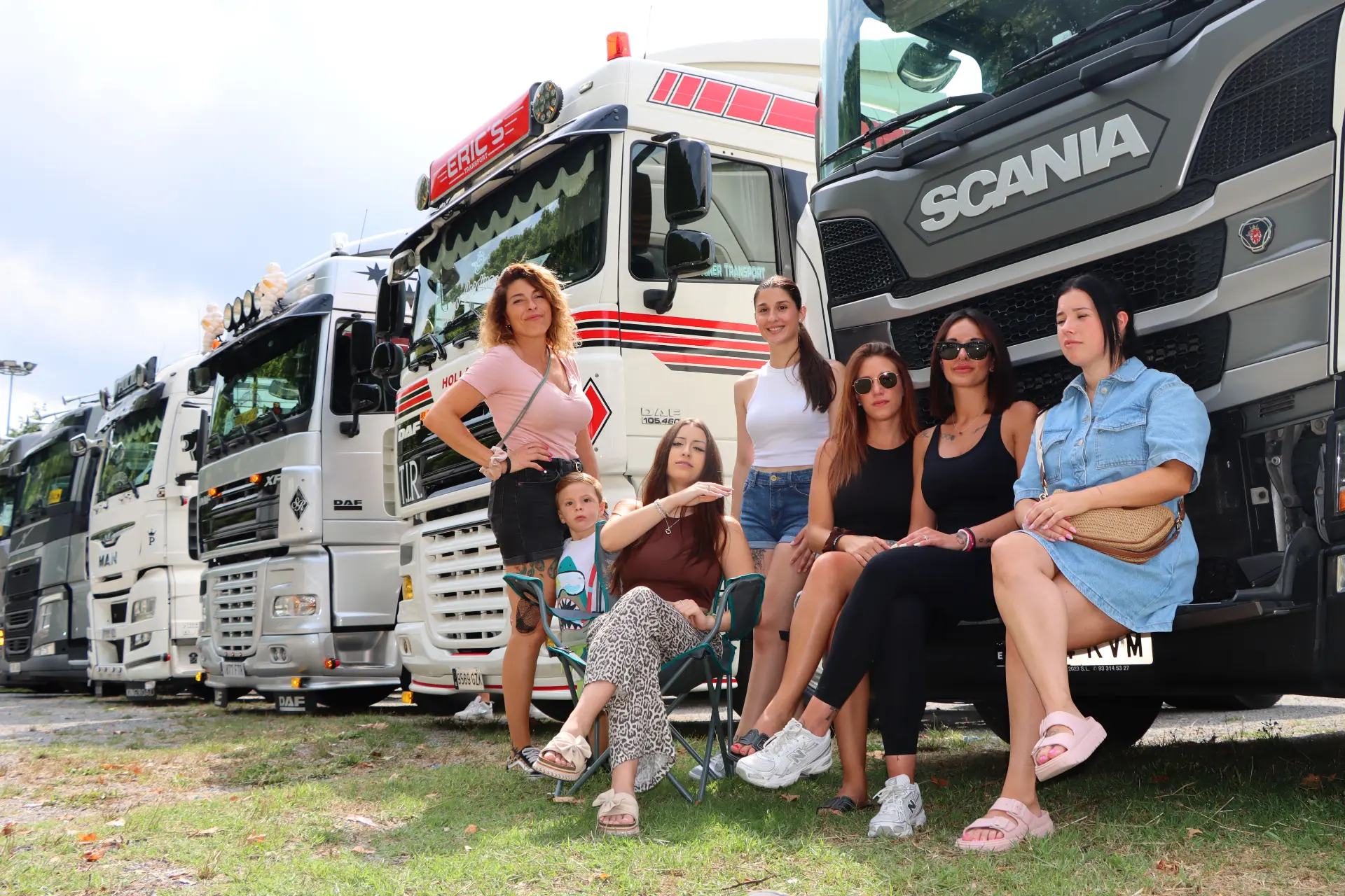 Siete mujeres y un niño posan sentados delante de camiones de Olot en un día soleado, disfrutando de la concentración de camiones.