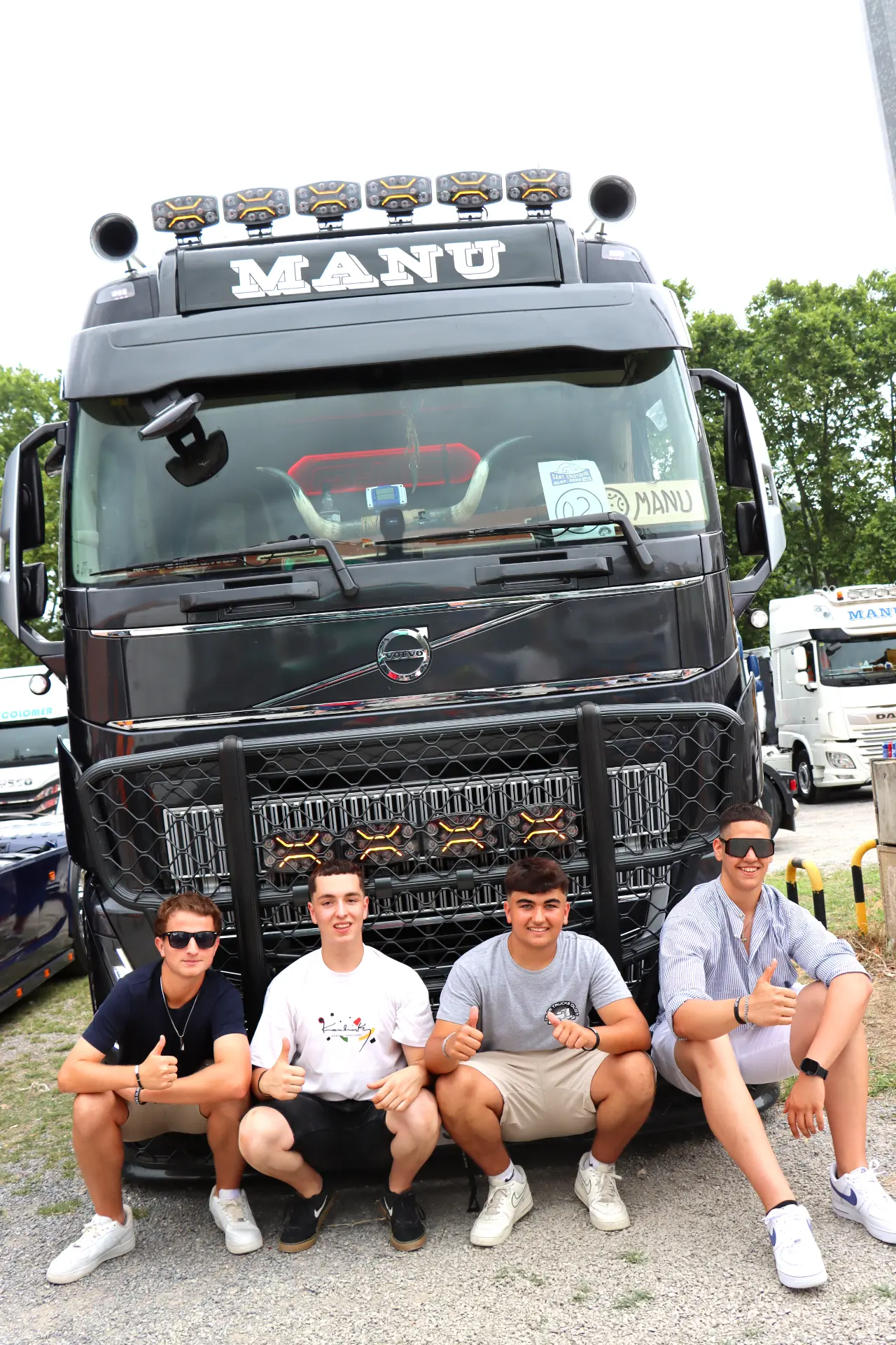 Cuatro jóvenes posan y sonríen delante de un gran camión negro con la inscripción "MANU" en una concentración de camiones en Olot.