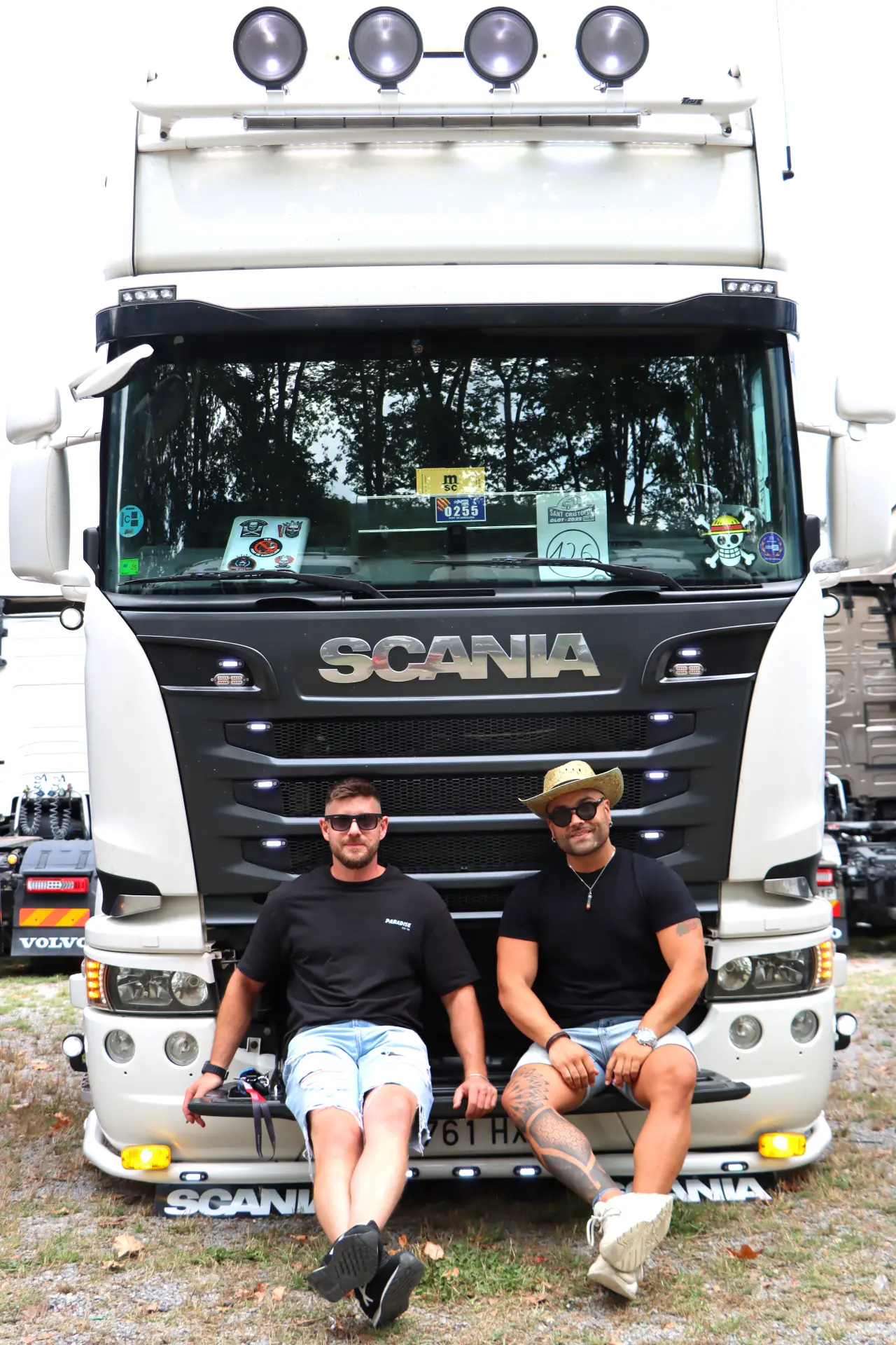 Dos hombres sentados en el parachoques de un camión Scania blanco sonríen a la cámara durante la concentración de camiones en Olot.