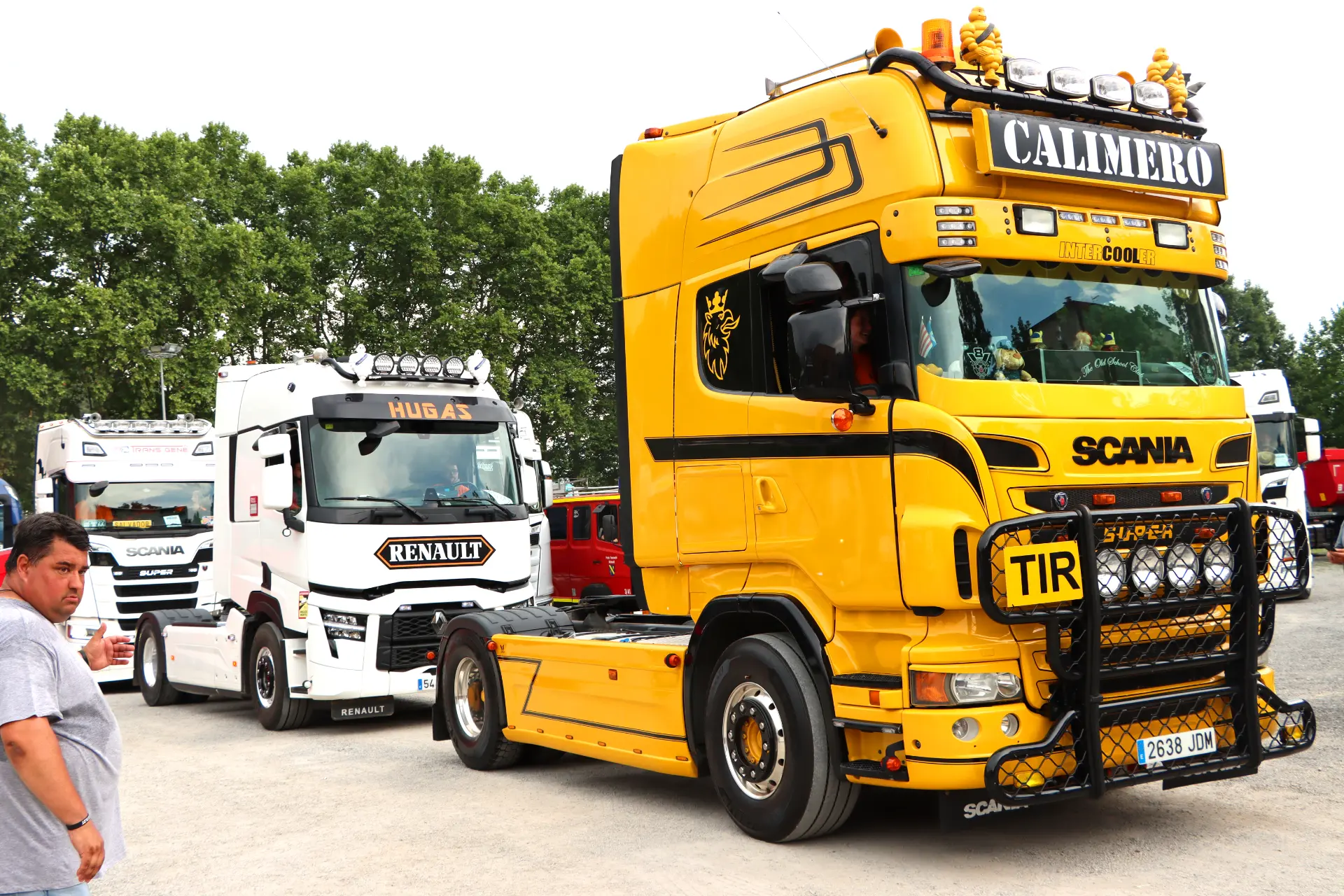Un camión Scania amarillo brillante con la etiqueta "CALMERO" está aparcado junto a un camión Renault blanco en una concentración de camiones en Olot; un hombre camina cerca.