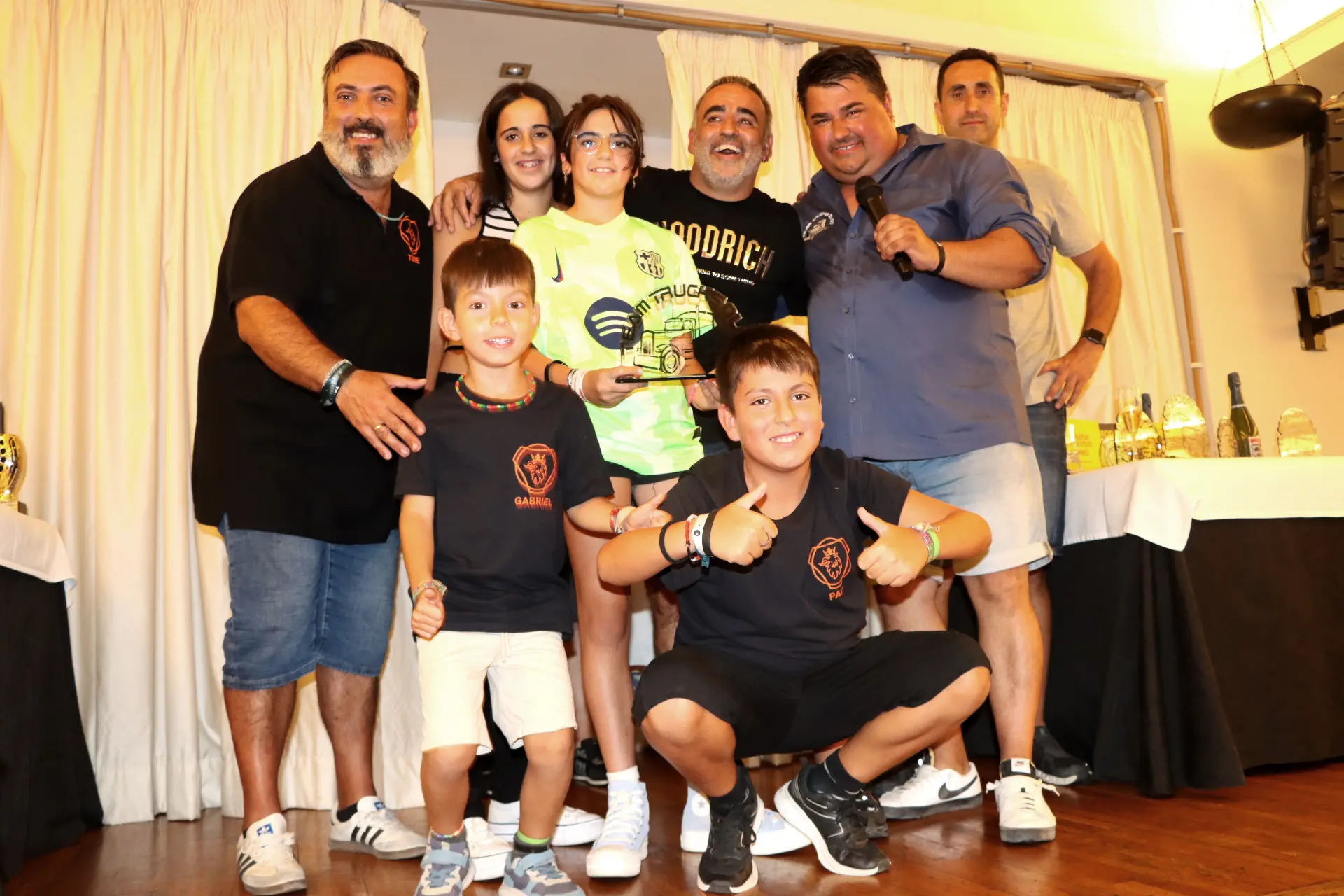 Grupo de personas sonrientes, incluidos niños, posando en el interior con un trofeo delante de cortinas blancas, celebrando la XI Concentración de camiones de Olot.