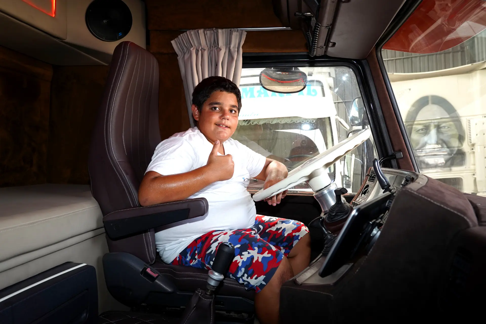 Un niño con camisa blanca levanta el pulgar sentado en el asiento del conductor de un camión, junto a una pegatina en la que se lee "Borrador automático".