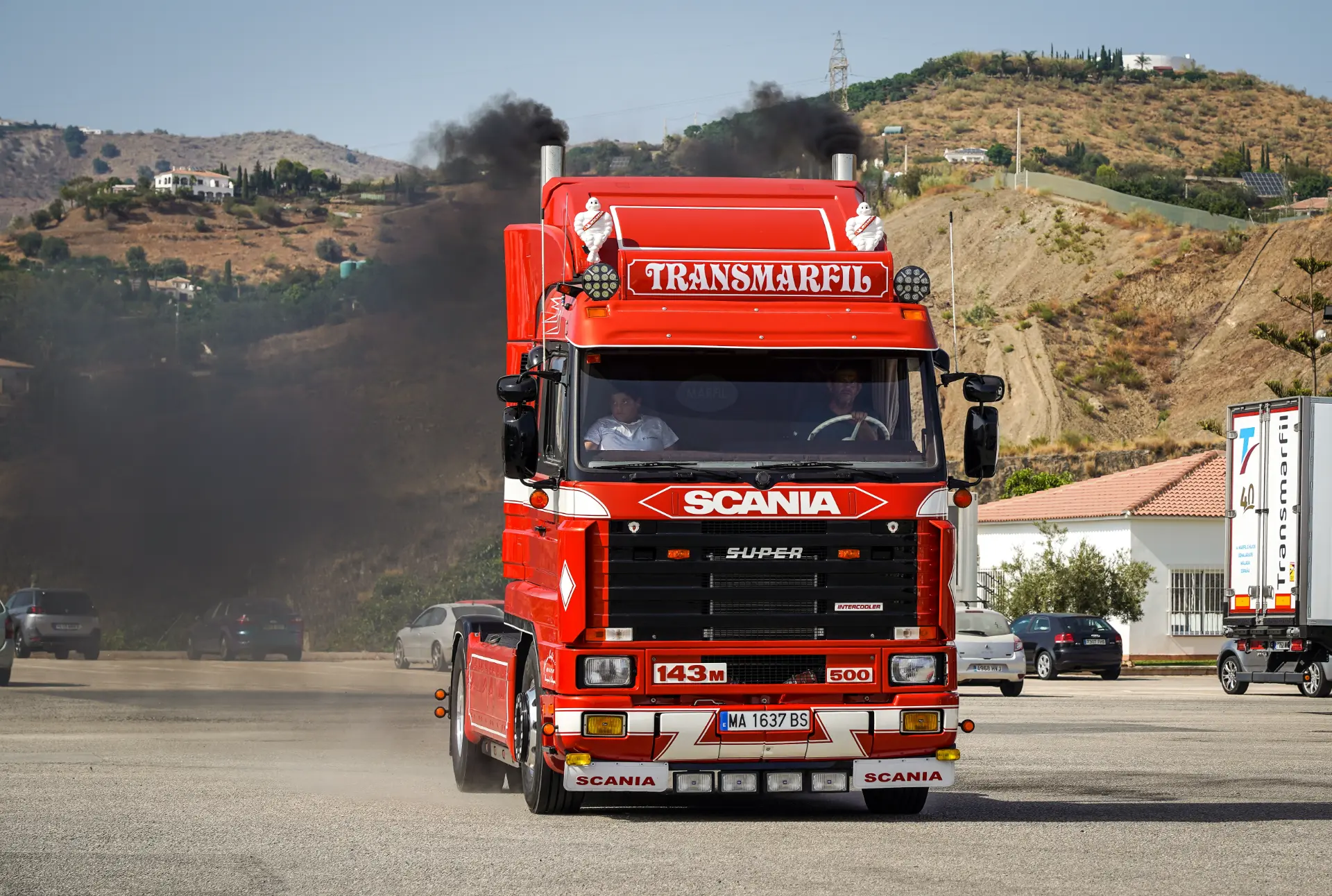 Un camión Scania rojo con borrador automático emite humo negro por el tubo de escape mientras circula por un aparcamiento.