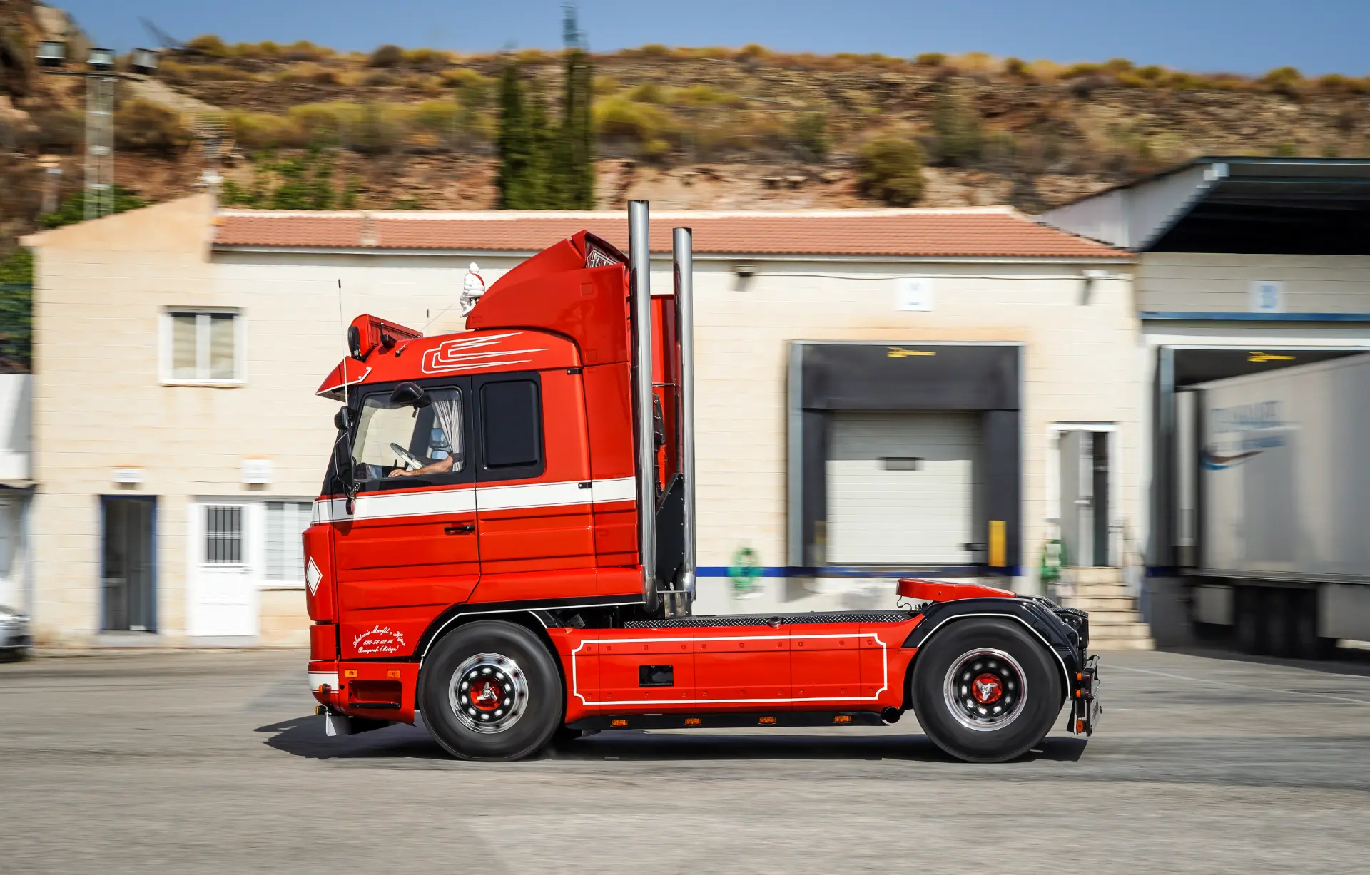 Una cabina roja de semirremolque, con sistema de borrador automático, pasa por delante de una nave industrial sin su remolque.