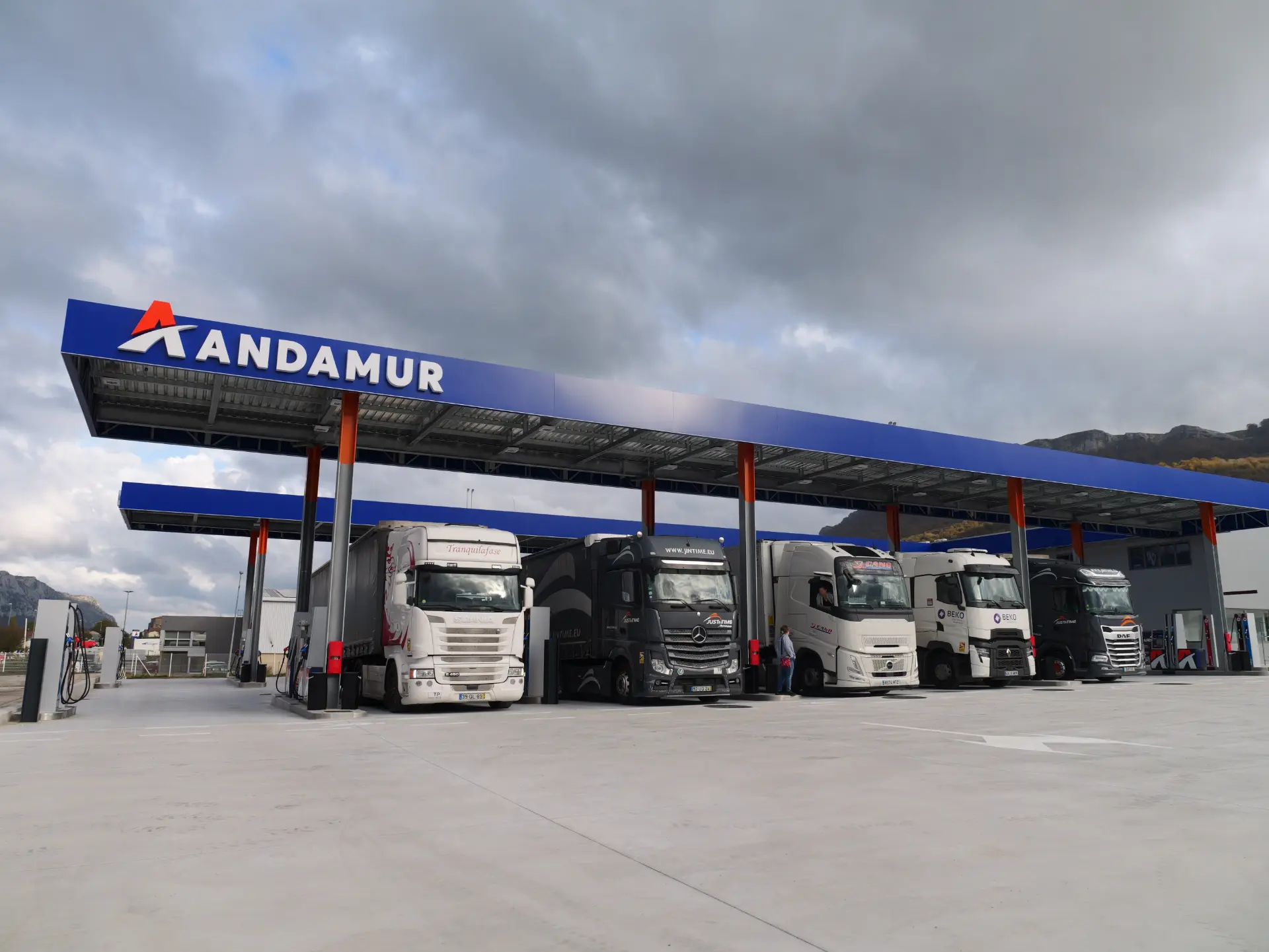 Cinco grandes camiones aparcados bajo la marquesina azul de una gasolinera de Andamur en un día nublado, aprovechando las áreas de descanso diseñadas con medidas reales para la comodidad de los conductores.