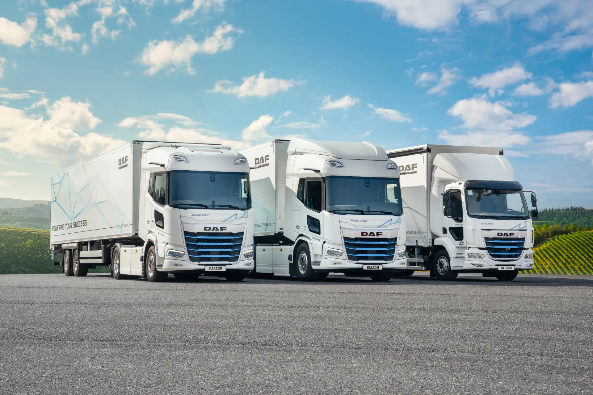 Tres camiones eléctricos DAF blancos, que representan la innovación Cero Emisiones de DAF Trucks España, están aparcados uno al lado del otro en una superficie pavimentada con un fondo escénico.