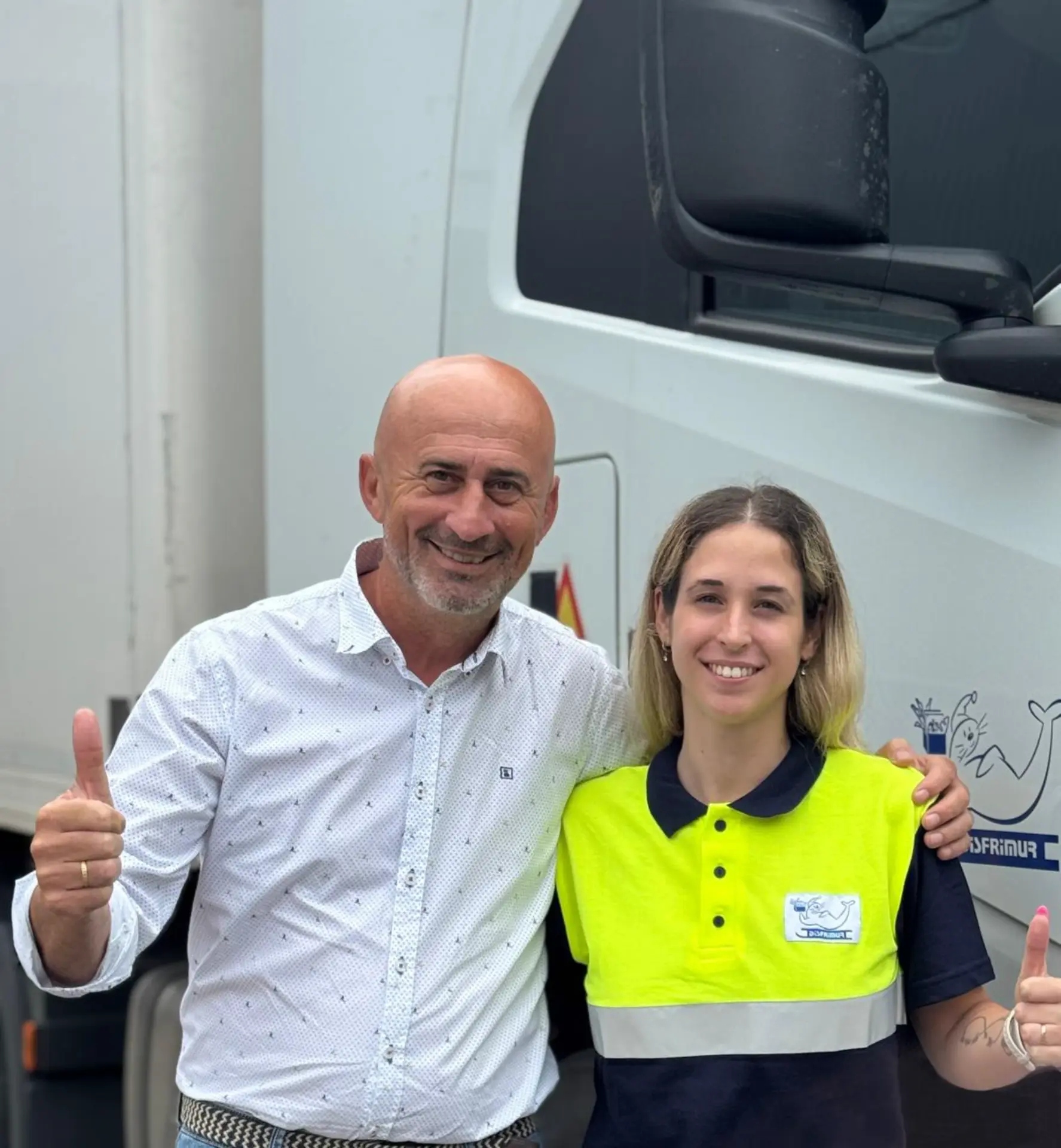 Dos personas sonriendo y dando pulgares arriba delante de un camión blanco, representando con orgullo a las mujeres camioneras y el espíritu de soy camionera con disfrimur.