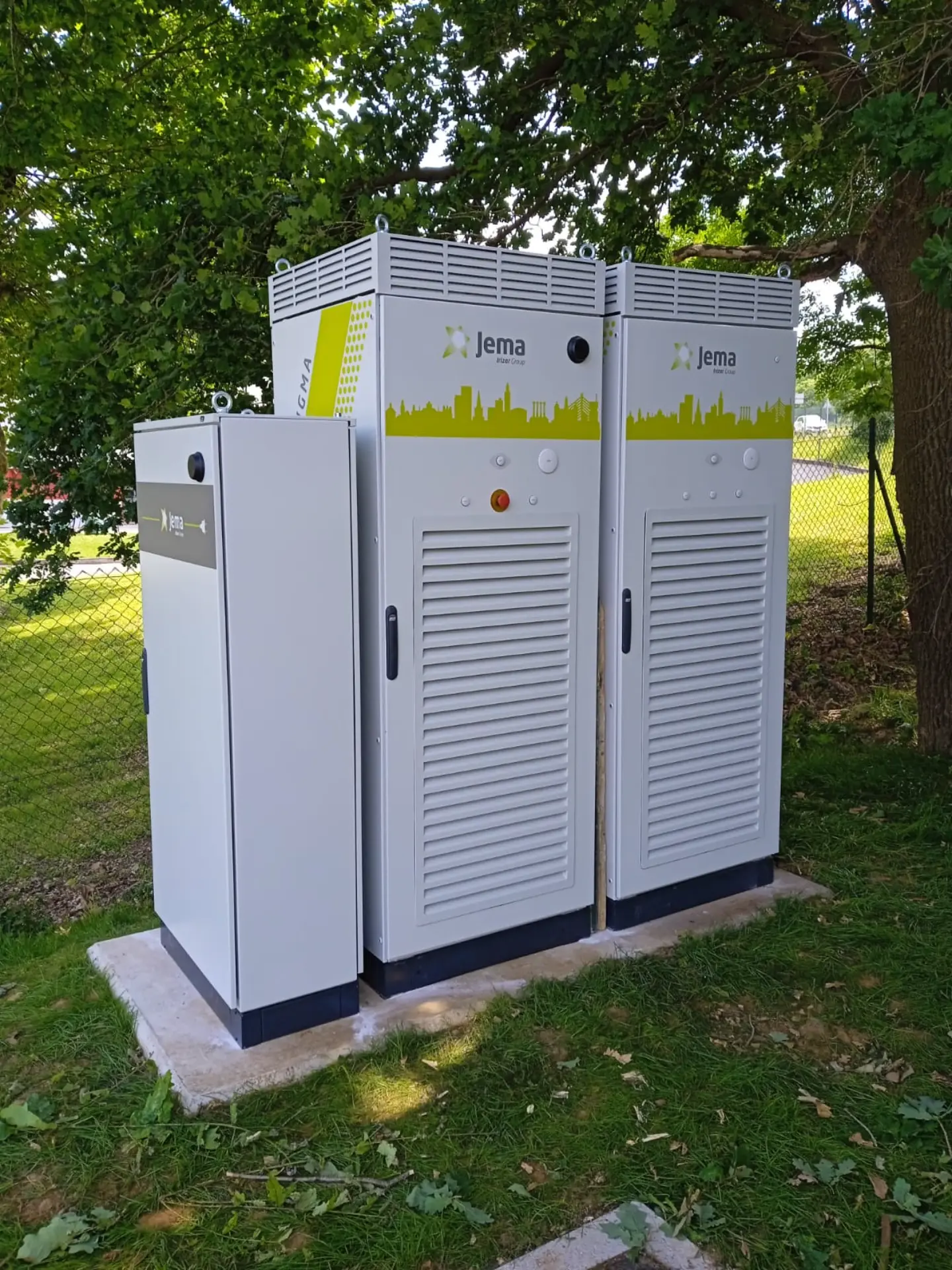 Dos grandes armarios eléctricos blancos de exterior con la etiqueta "Jema Energy" se alzan sobre hormigón bajo los árboles junto a una valla, listos para adaptarse a cualquier necesidad.