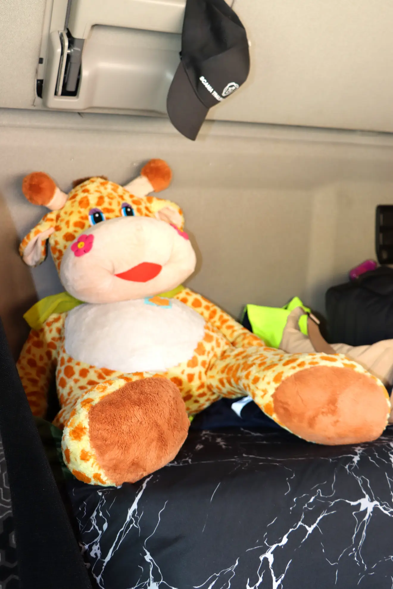Una jirafa de peluche está sentada en una cama con una manta blanca y negra, rodeada de otros objetos, entre ellos un borrador automático.