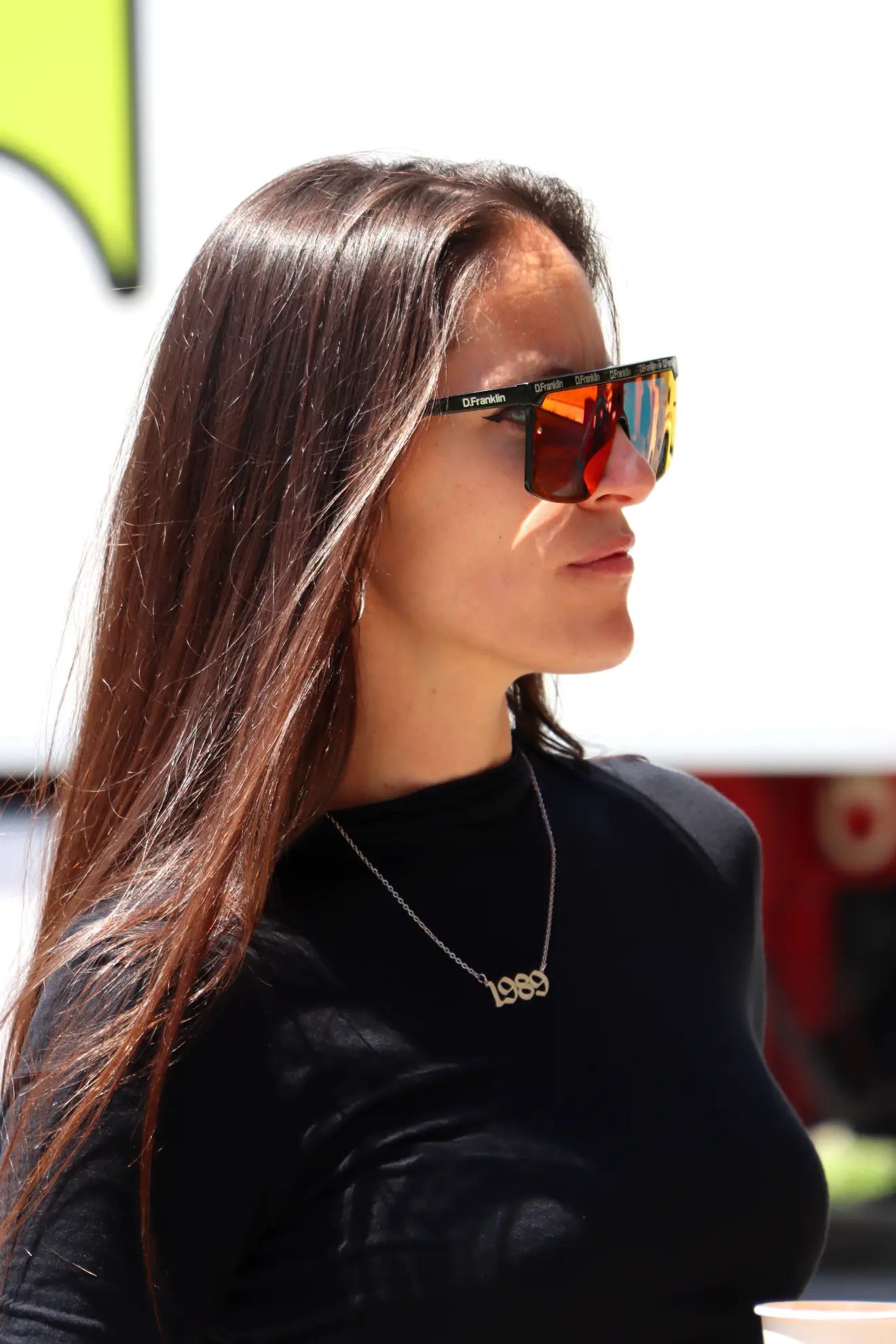 Mujer de pelo largo y castaño, con gafas de sol, top negro y collar "1999", de pie al aire libre, con un look chic, casi como si la hubiera peinado un Borrador automático.