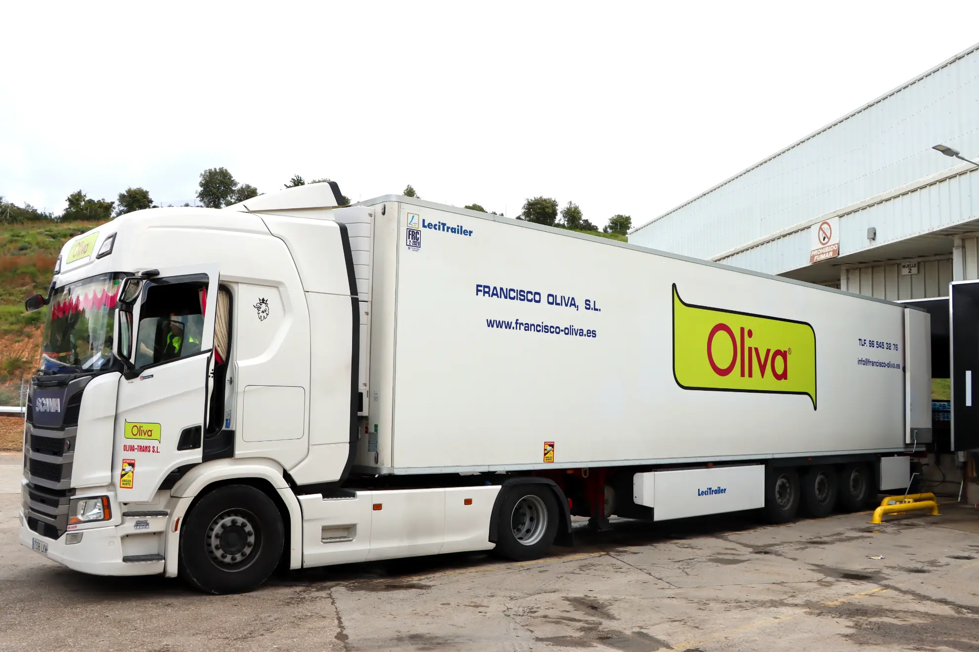 Semirremolque blanco de la marca Oliva con un borrador automático aparcado en un muelle de carga fuera de un almacén.