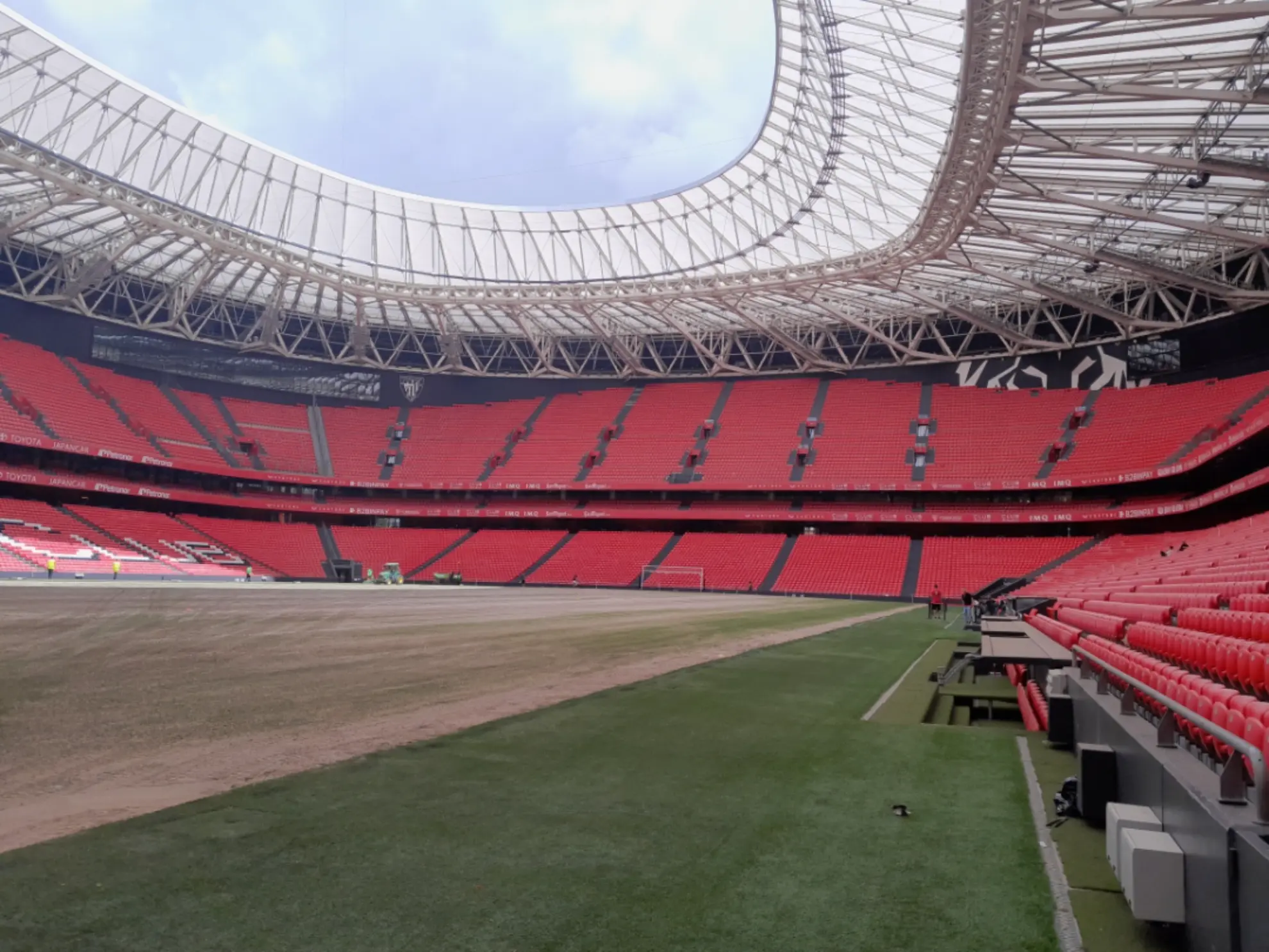 Estadio vacío con asientos rojos y un campo parcialmente desnudo, visto desde el nivel del terreno de juego cerca de las gradas.