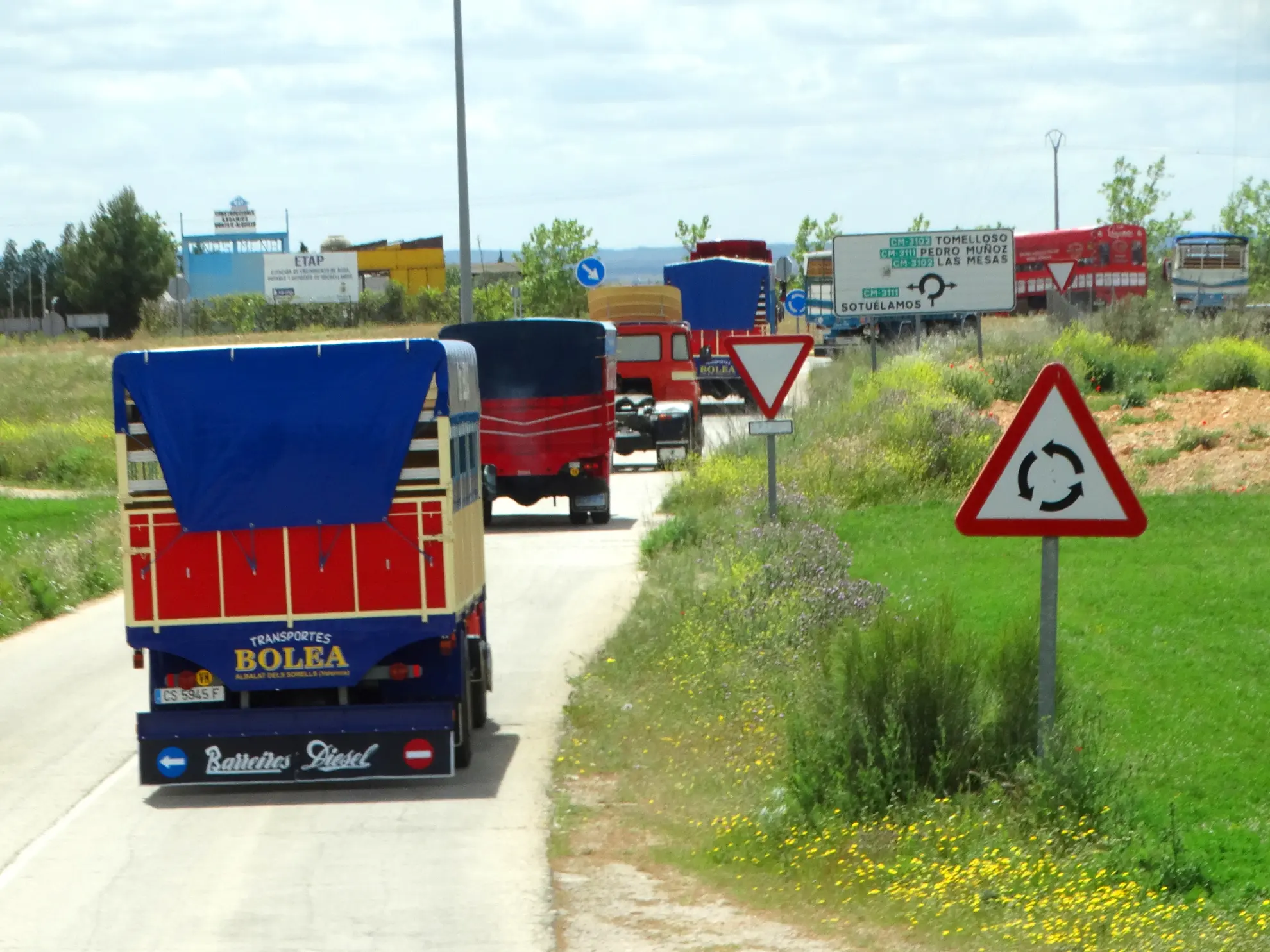 Camiones de colores circulan por una carretera rural cerca de una rotonda, con señales de tráfico, vegetación y un aviso de Borrador automático a la vista.