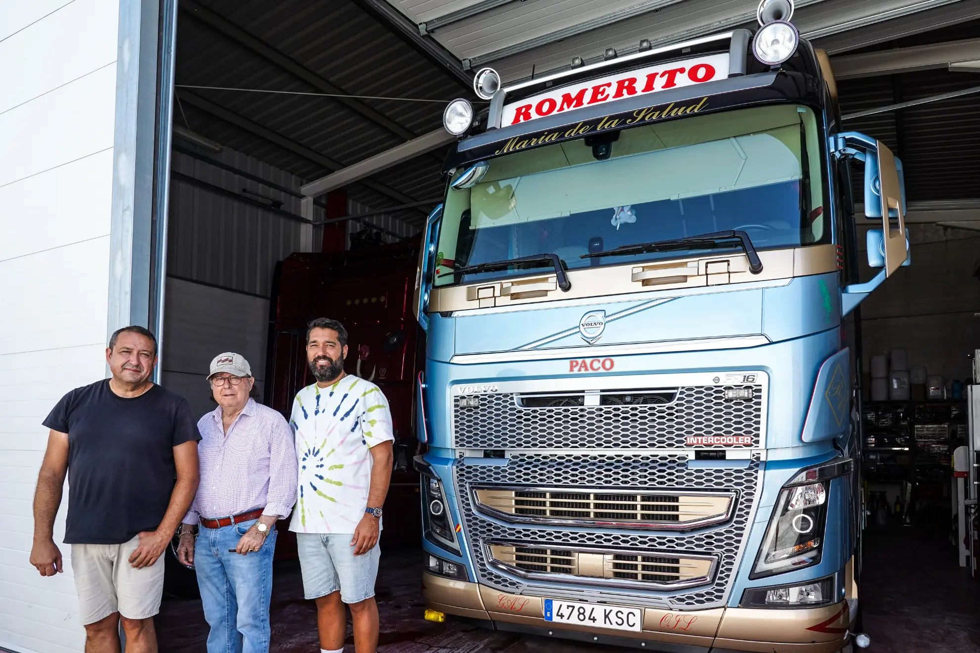 Tres hombres permanecen de pie junto a un gran camión azul rotulado "ROMERITO" en un garaje, discutiendo detalles cerca de un borrador automático.