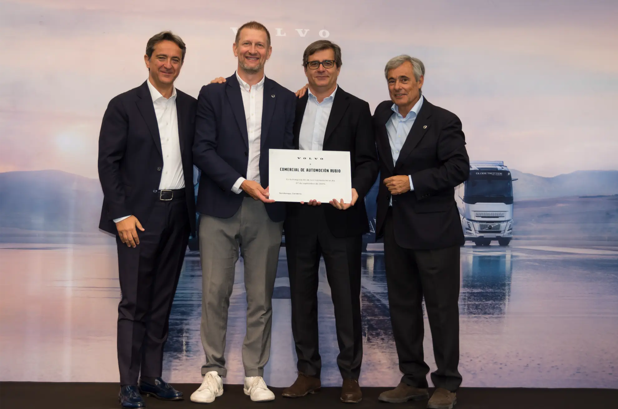 Cuatro hombres trajeados de pie, juntos y sosteniendo un certificado, con camiones y agua al fondo, celebrando el éxito de la implantación de un sistema de borrador automático.
