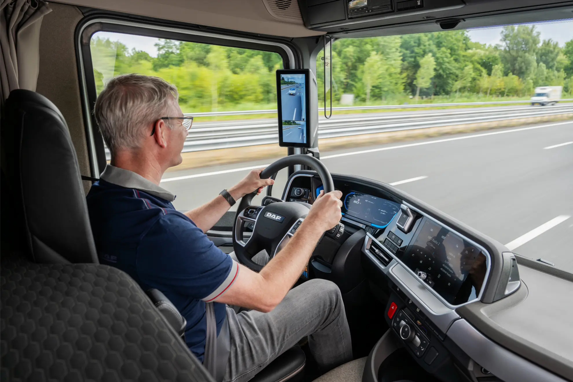 Hombre conduciendo un moderno camión DAF eléctrico por la autopista, utilizando un salpicadero digital y la pantalla de la cámara del retrovisor lateral, destacando las avanzadas características y la notable autonomía que DAF eléctrico ofrece para los viajes de larga distancia.