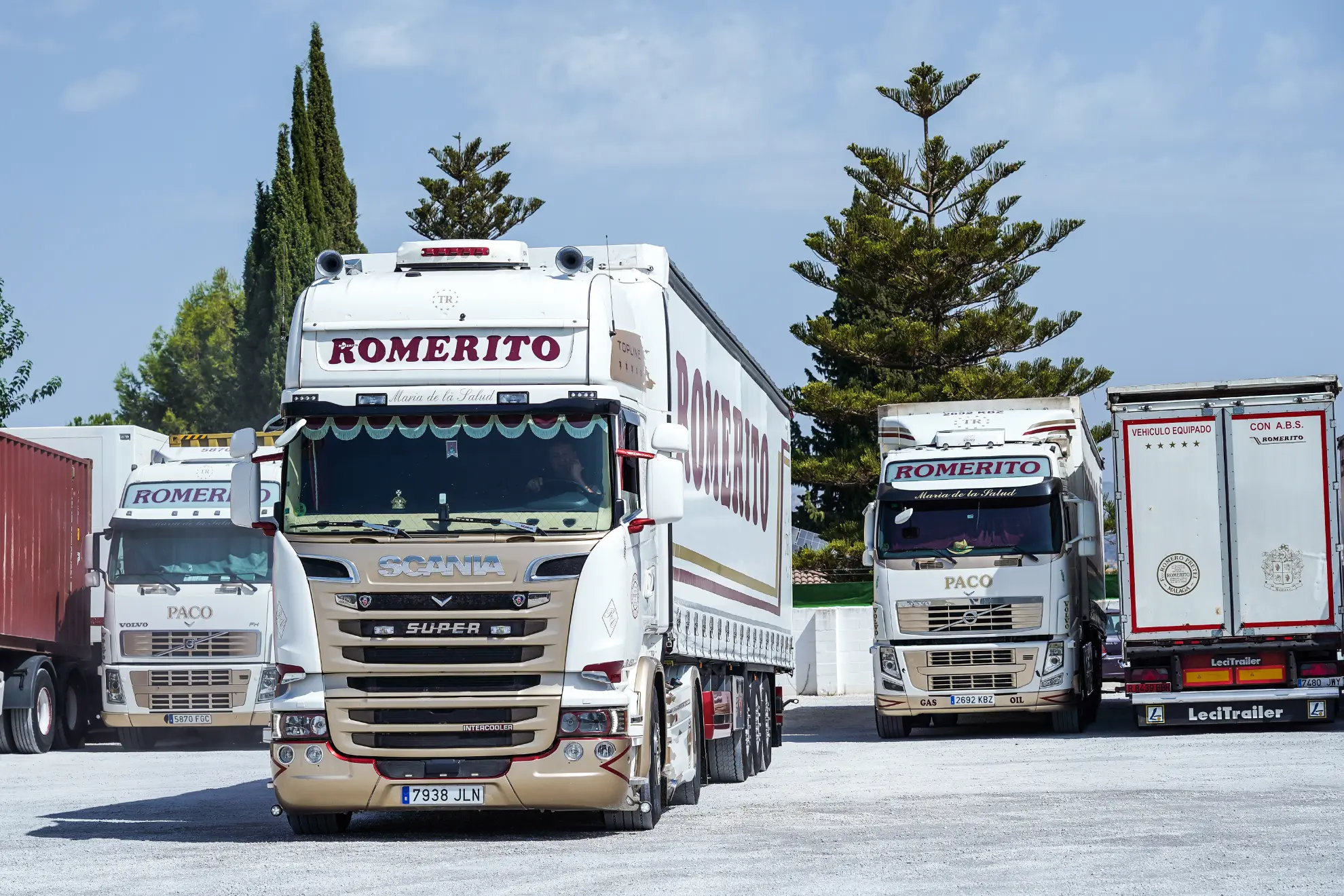 Tres grandes camiones de transporte blancos rotulados "ROMERITO", con tecnología de borrador automático, están aparcados en un solar con árboles al fondo.