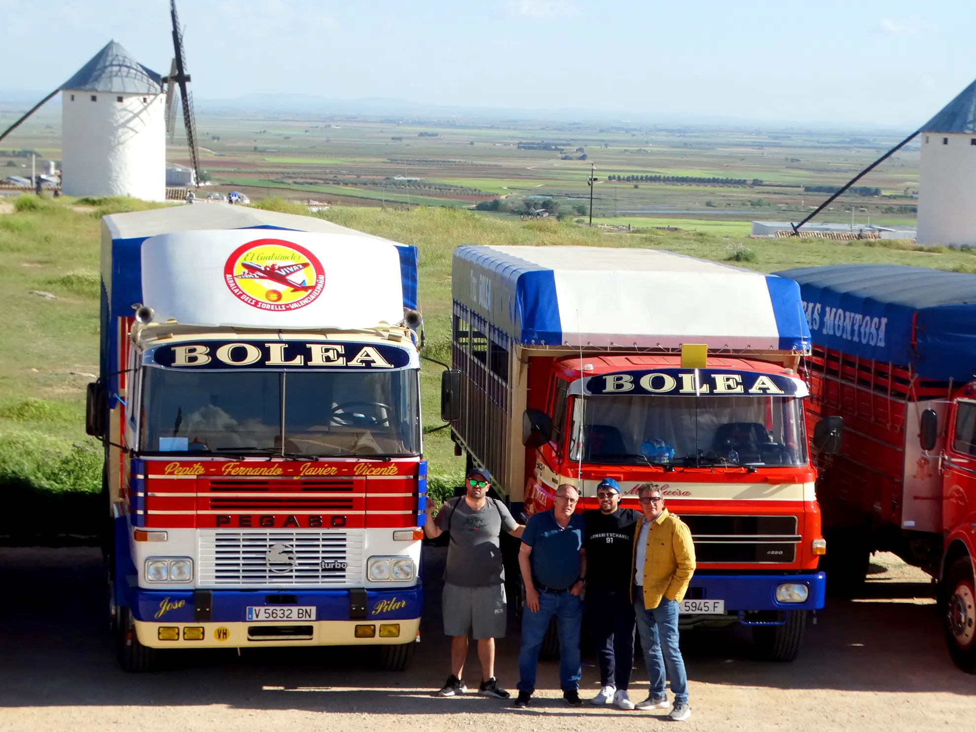 Cuatro hombres de pie delante de dos coloridos camiones Bolea, con molinos de viento y campos al fondo, mientras un borrador automático zumba tranquilamente cerca.