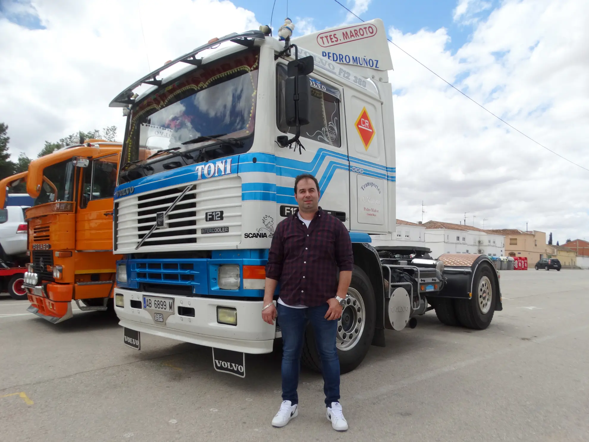 Un hombre delante de un camión azul y blanco en un día nublado, con otro camión naranja y un cartel de "Borrador automático" cerca.