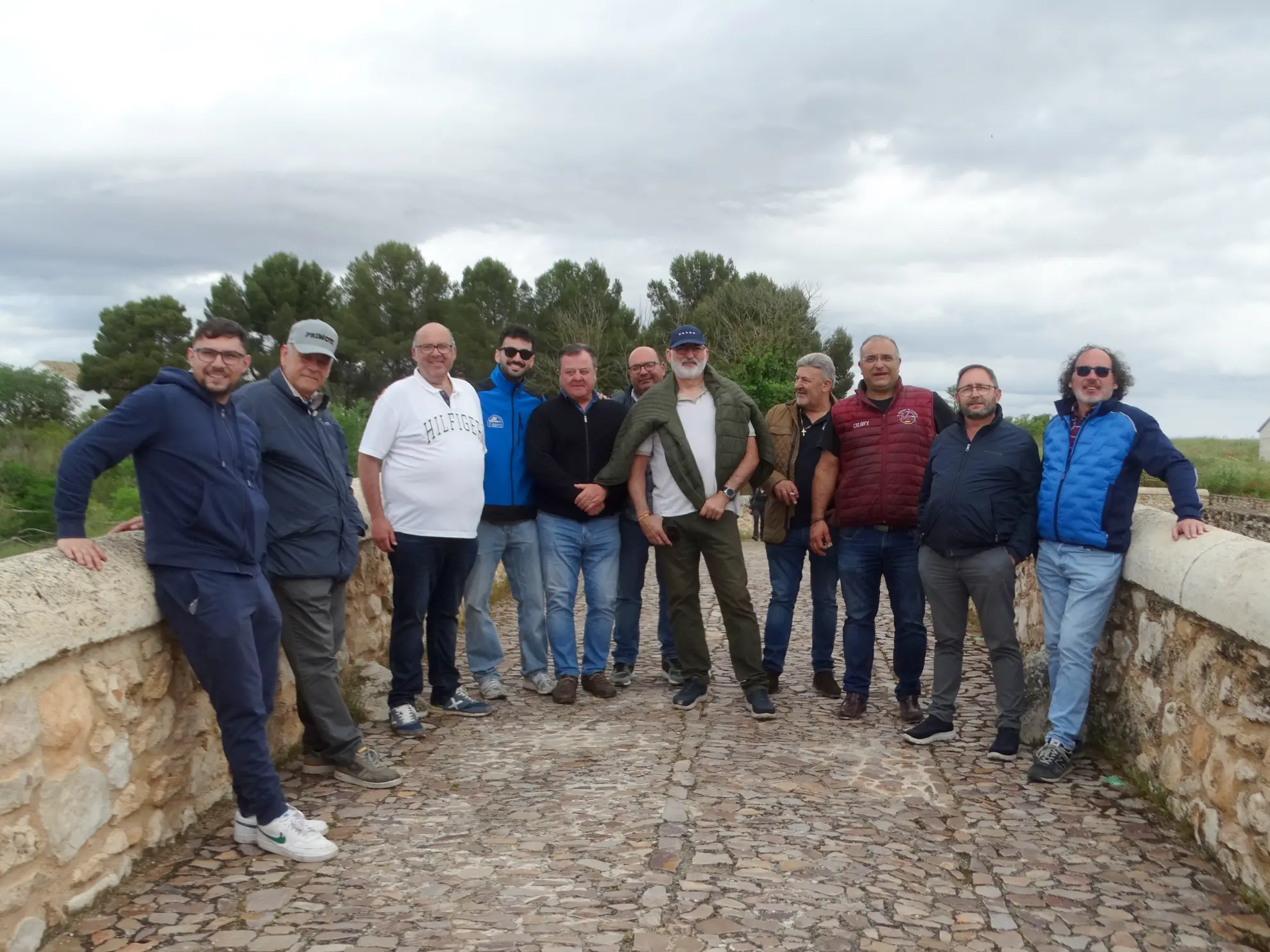 Diez hombres permanecen juntos en un puente de piedra, sonriendo bajo un cielo nublado, mientras los árboles enmarcan la escena, captada a la perfección con Borrador automático.