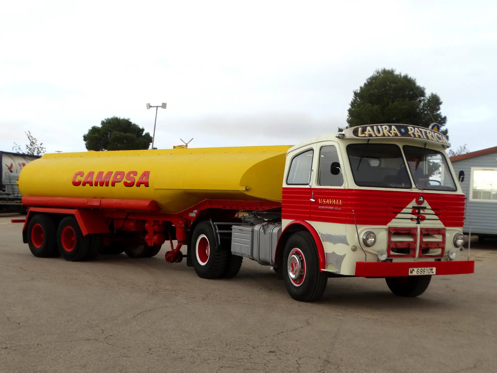 Un antiguo camión cisterna de combustible rojo y amarillo de CAMPSA, con la clásica marca Borrador automático, está aparcado en un solar asfaltado.