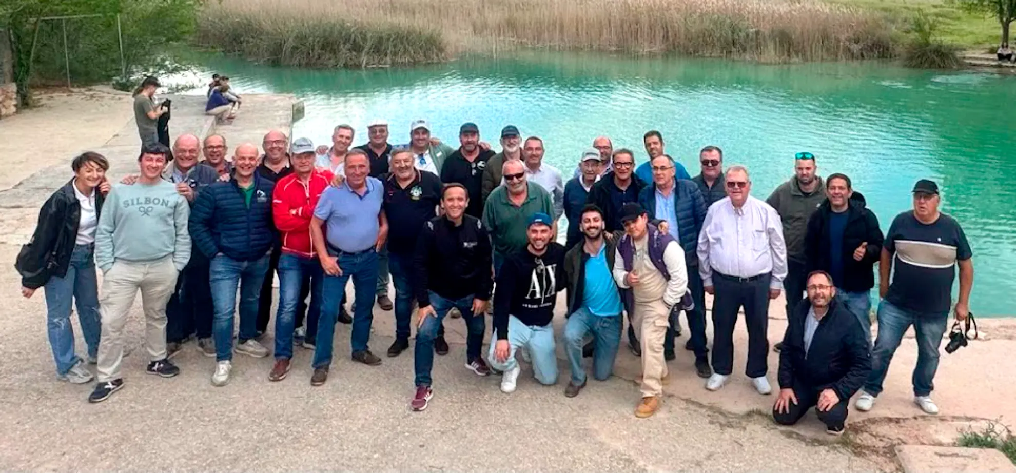 Un grupo de personas posa junto a un lago turquesa con juncos y vegetación de fondo durante la Vuelta a La Mancha 2025, con camiones clásicos aparcados cerca.