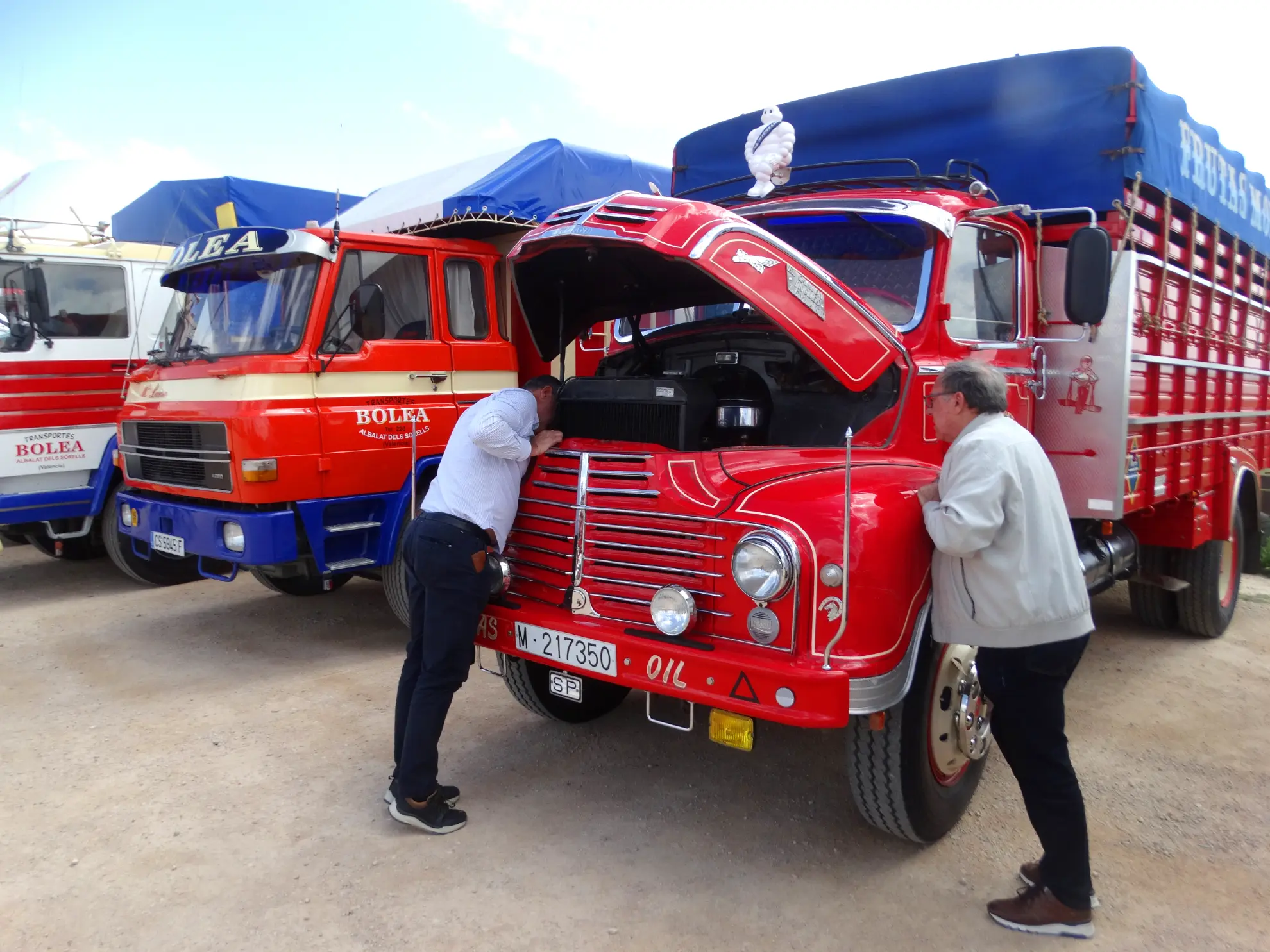 Dos personas inspeccionan el motor de un clásico camión rojo con el capó abierto, aparcado entre otros coloridos camiones durante la Vuelta a La Mancha 2025.