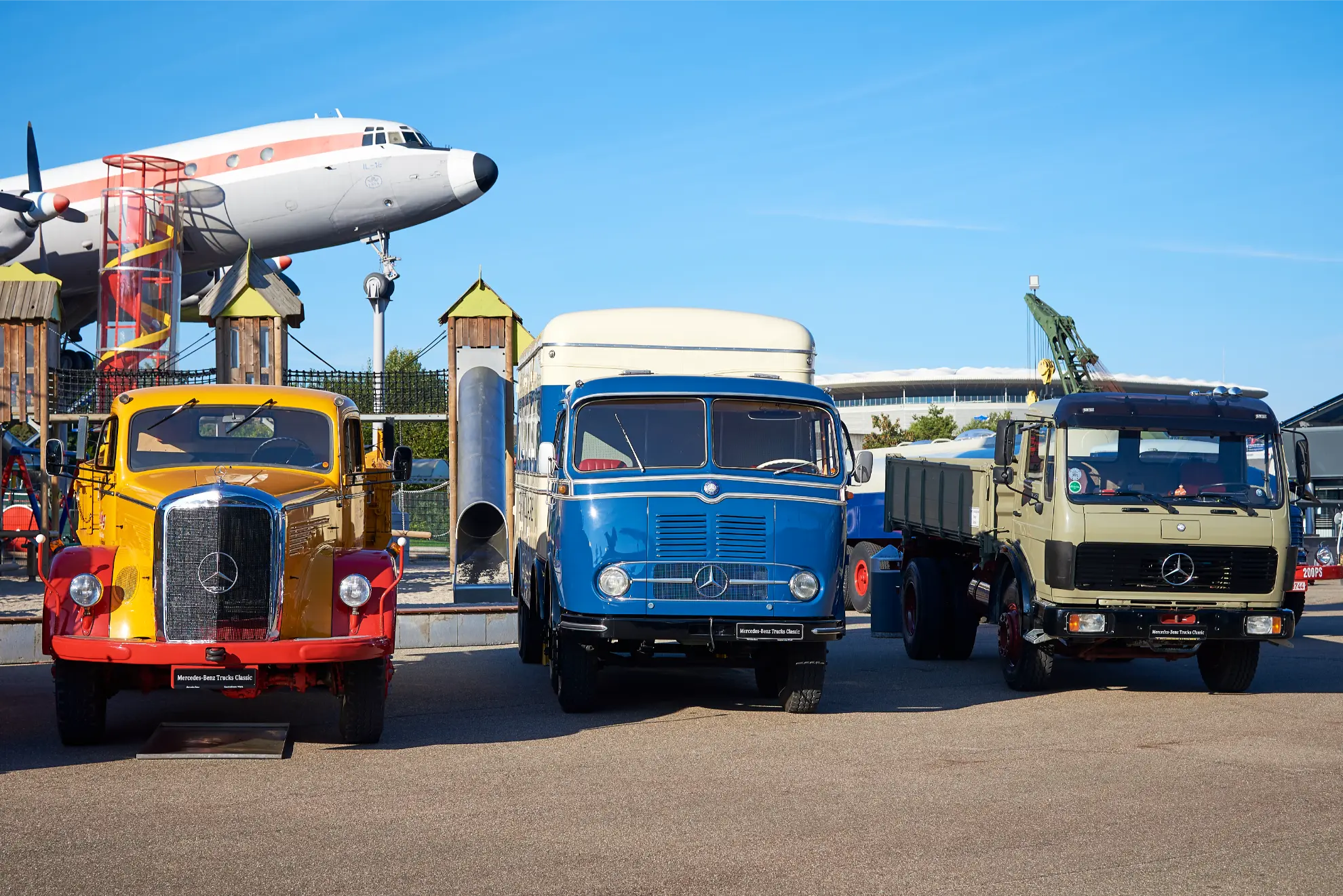 Tres camiones Mercedes-Benz de época están aparcados uno al lado del otro, con un viejo avión al fondo, creando una escena que parece captada con un borrador automático.