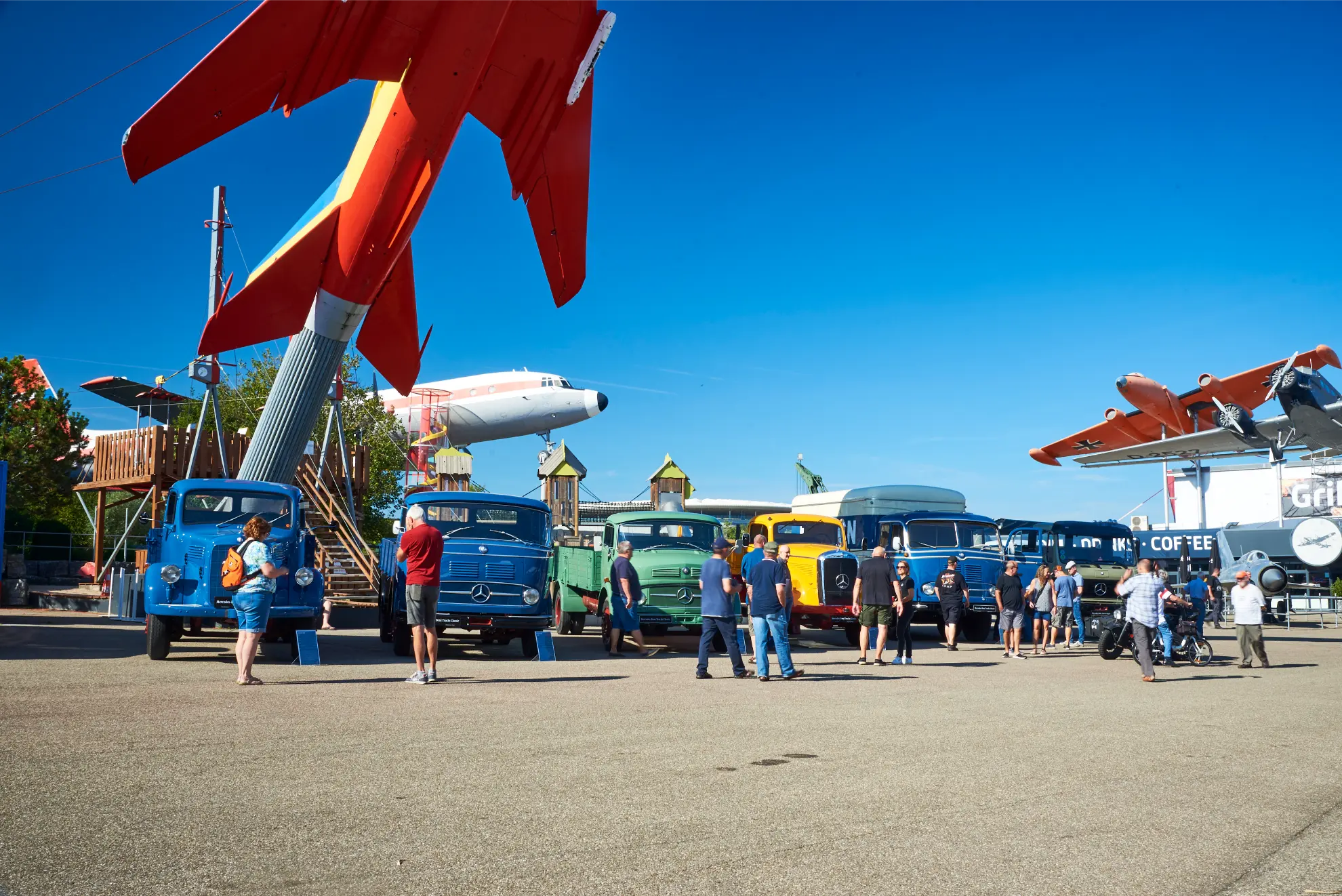Los visitantes admiran camiones históricos en una exposición al aire libre en Sinsheim, con Mercedes-Benz Trucks Classic en exhibición y aviones volando por encima contra un cielo azul claro.