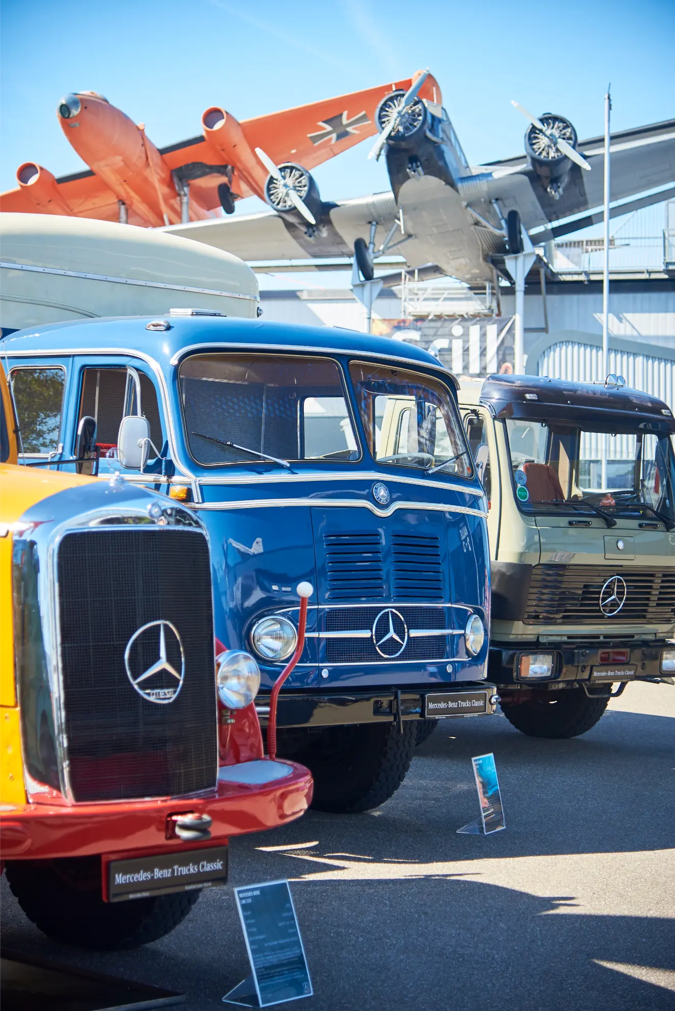 Camiones Mercedes-Benz clásicos Camiones históricos aparcados en fila en Sinsheim, con aviones de época expuestos sobre ellos.