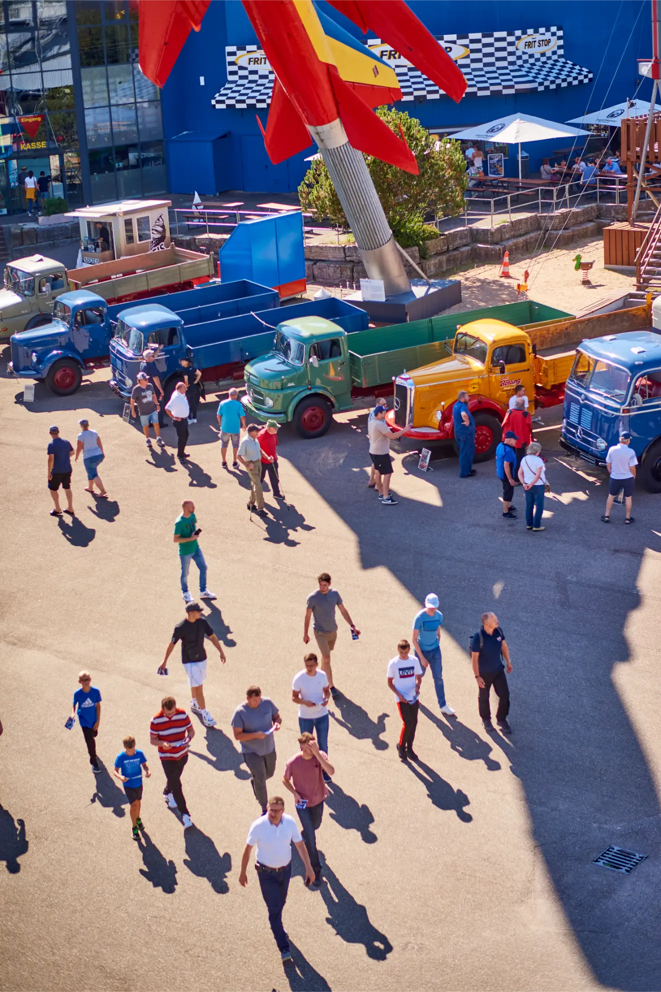 Gente paseando cerca de camiones históricos, entre ellos Mercedes-Benz Trucks Classic, en una exposición al aire libre en Sinsheim, con una gran estatua de un cohete encima proyectando una sombra.