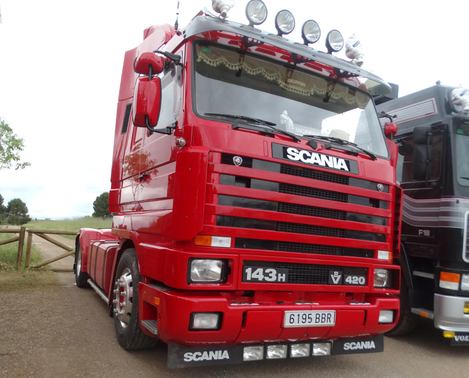 Camión Scania 143H 420 clásico rojo brillante aparcado al aire libre en tierra junto a otro camión, fotografiado de frente a la izquierda.