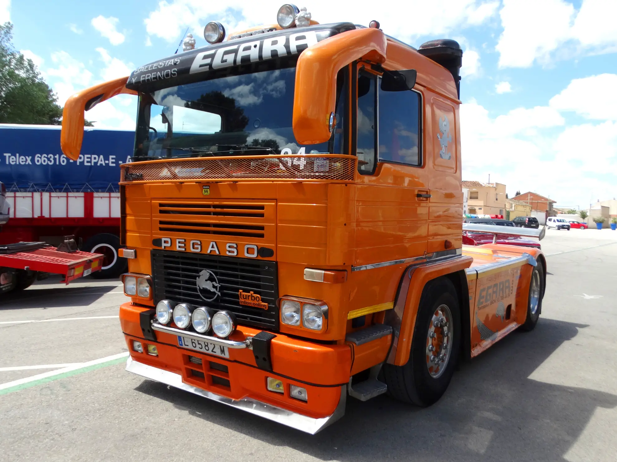 Camión Pegaso de color naranja brillante aparcado al aire libre en un día soleado, con edificios y otro camión al fondo, un vehículo clásico que suele verse apoyando eventos ciclistas como la Vuelta a La Mancha 2025.