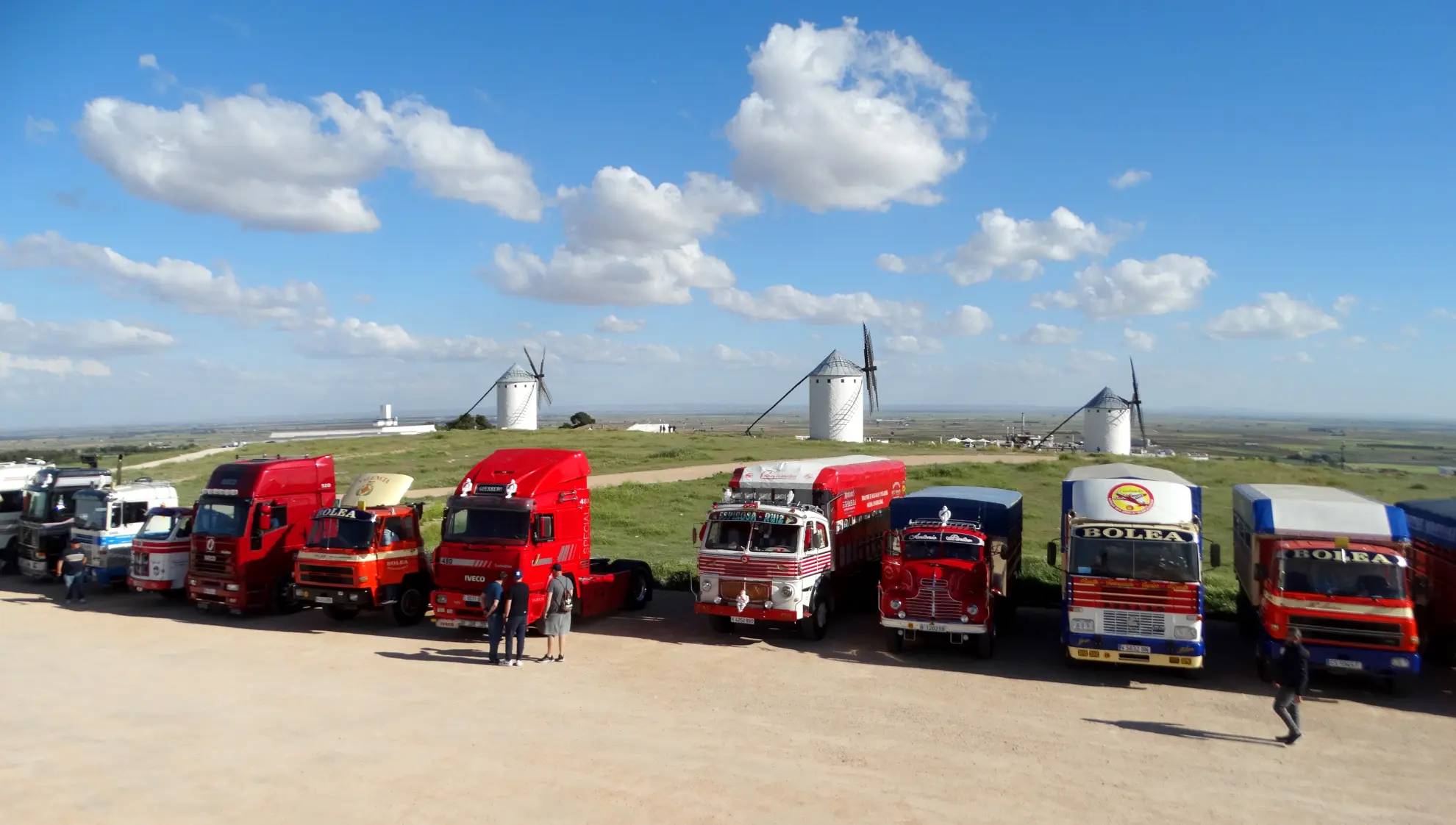Camiones clásicos aparcados en vibrante fila durante la Vuelta a La Mancha 2025, con molinos de viento y un cielo nublado creando un pintoresco telón de fondo.