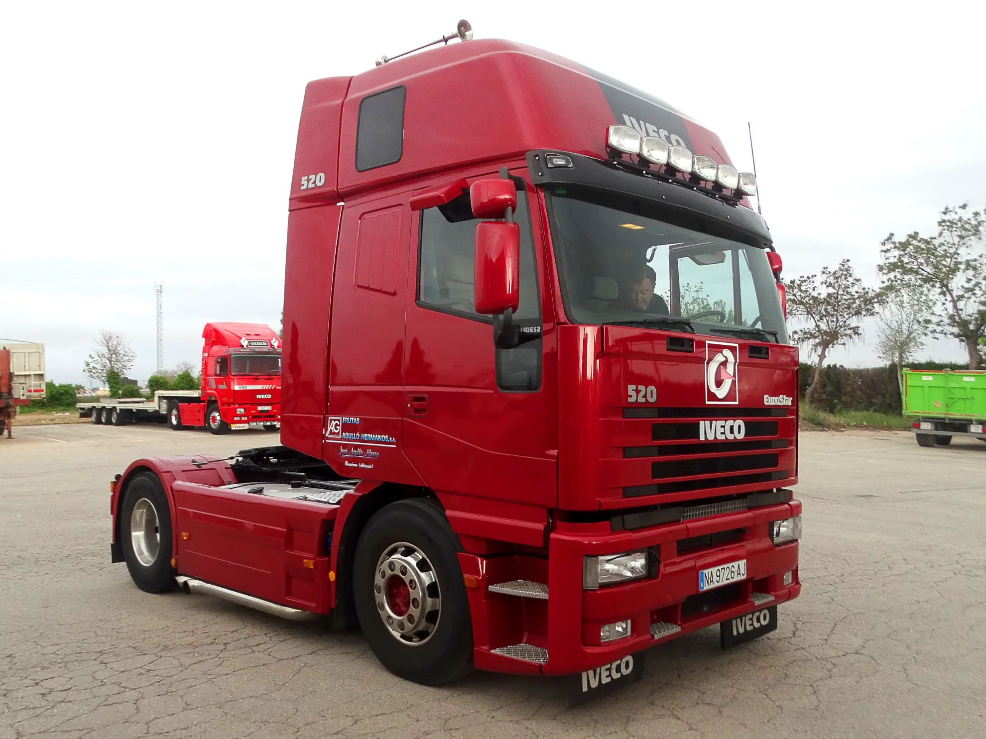 Un camión-cabina Iveco rojo, que forma parte de la concentración de camiones clásicos de la Vuelta a La Mancha 2025, está aparcado en un solar asfaltado con otros vehículos al fondo.