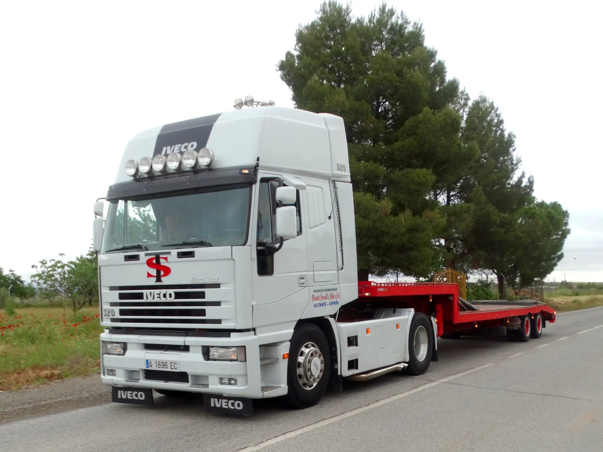 Semirremolque Iveco blanco con un remolque rojo de plataforma vacía aparcado al borde de una carretera cerca de árboles, listo para la Vuelta a La Mancha 2025: un ejemplo perfecto de camiones clásicos en acción.