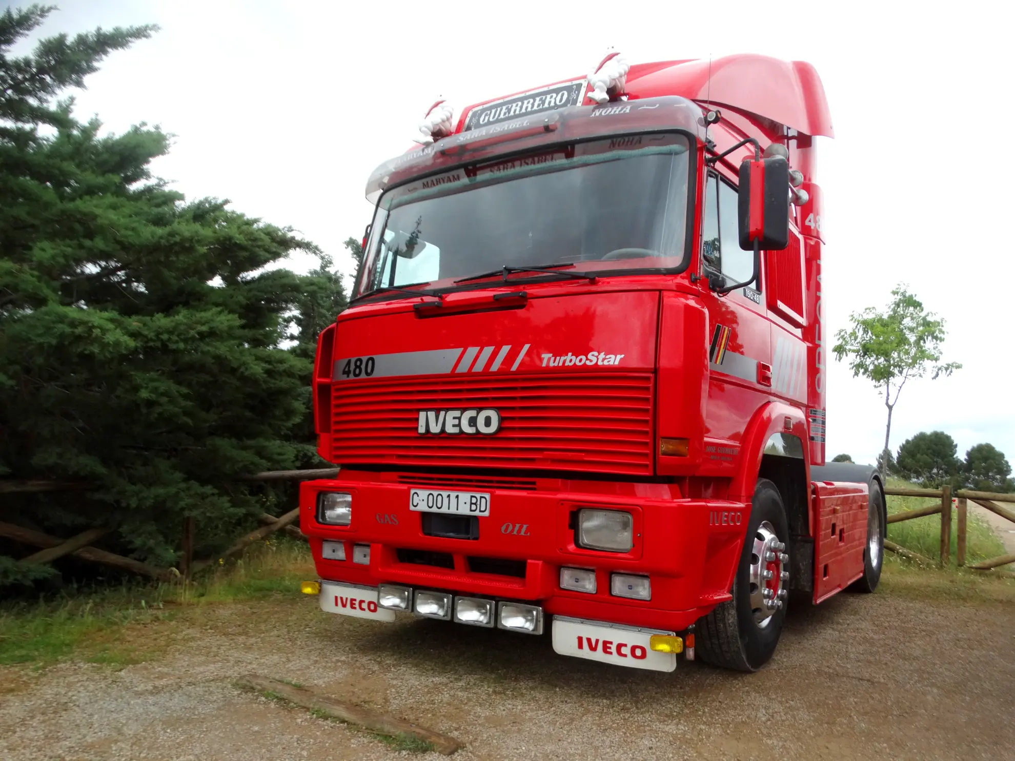 A red Iveco TurboStar, uno de los camiones clásicos, está estacionado en un camino de tierra cerca de árboles y una valla de madera durante la Vuelta a La Mancha 2025.