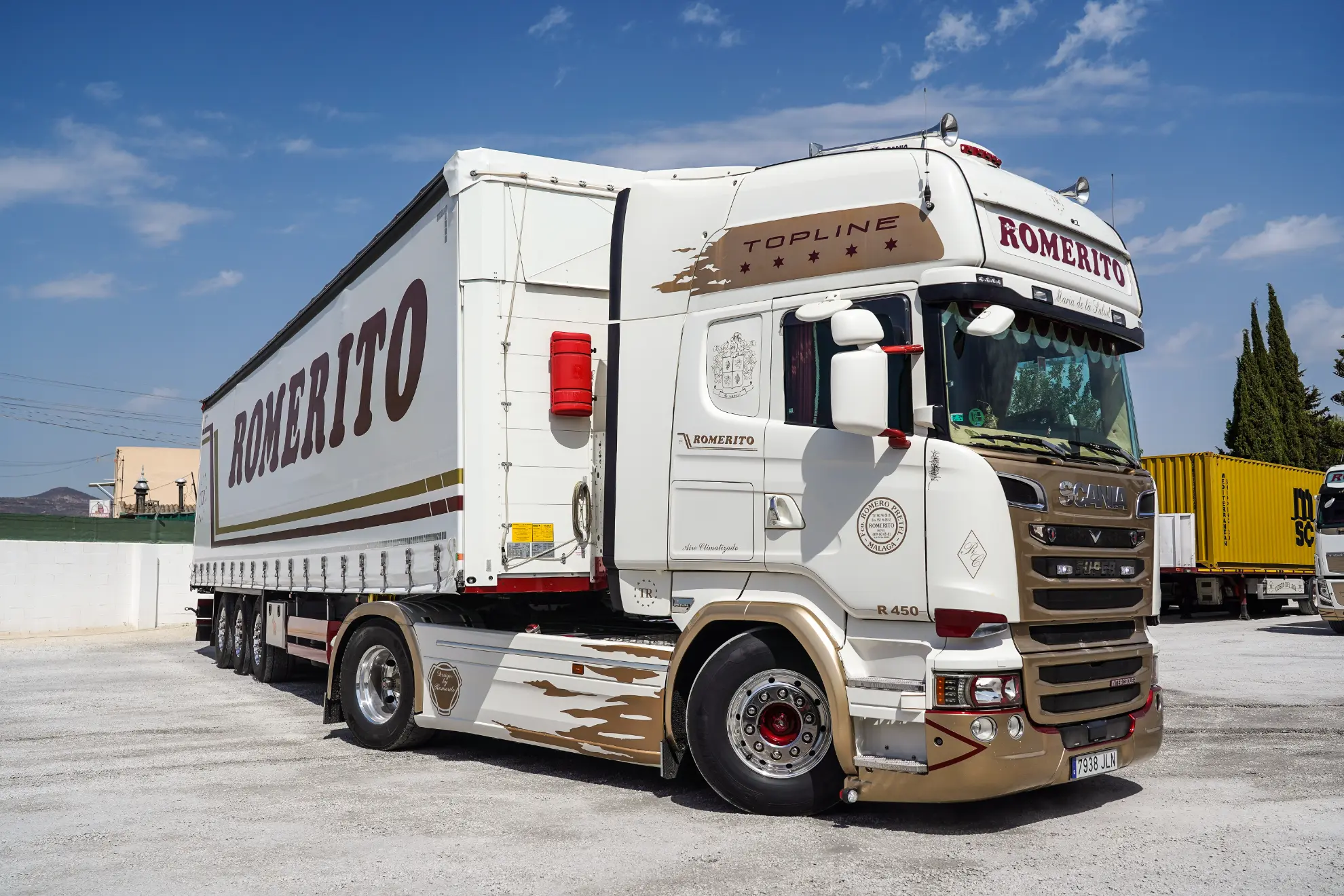 Camión Romerito blanco y dorado con remolque, con Borrador automático, aparcado en un solar en un día soleado.
