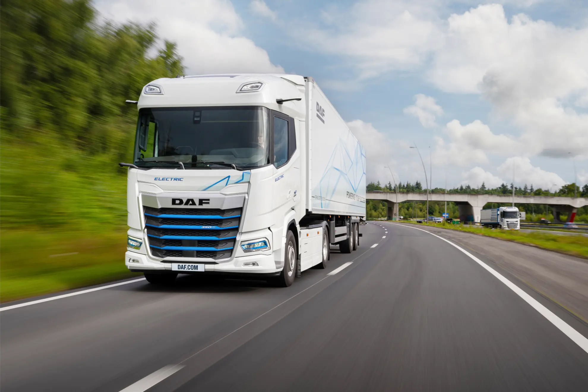 Camión eléctrico DAF blanco circulando por una autopista con árboles y un puente de fondo, mostrando la autonomía DAF eléctrica y los beneficios en TCO de los camiones DAF eléctricos.