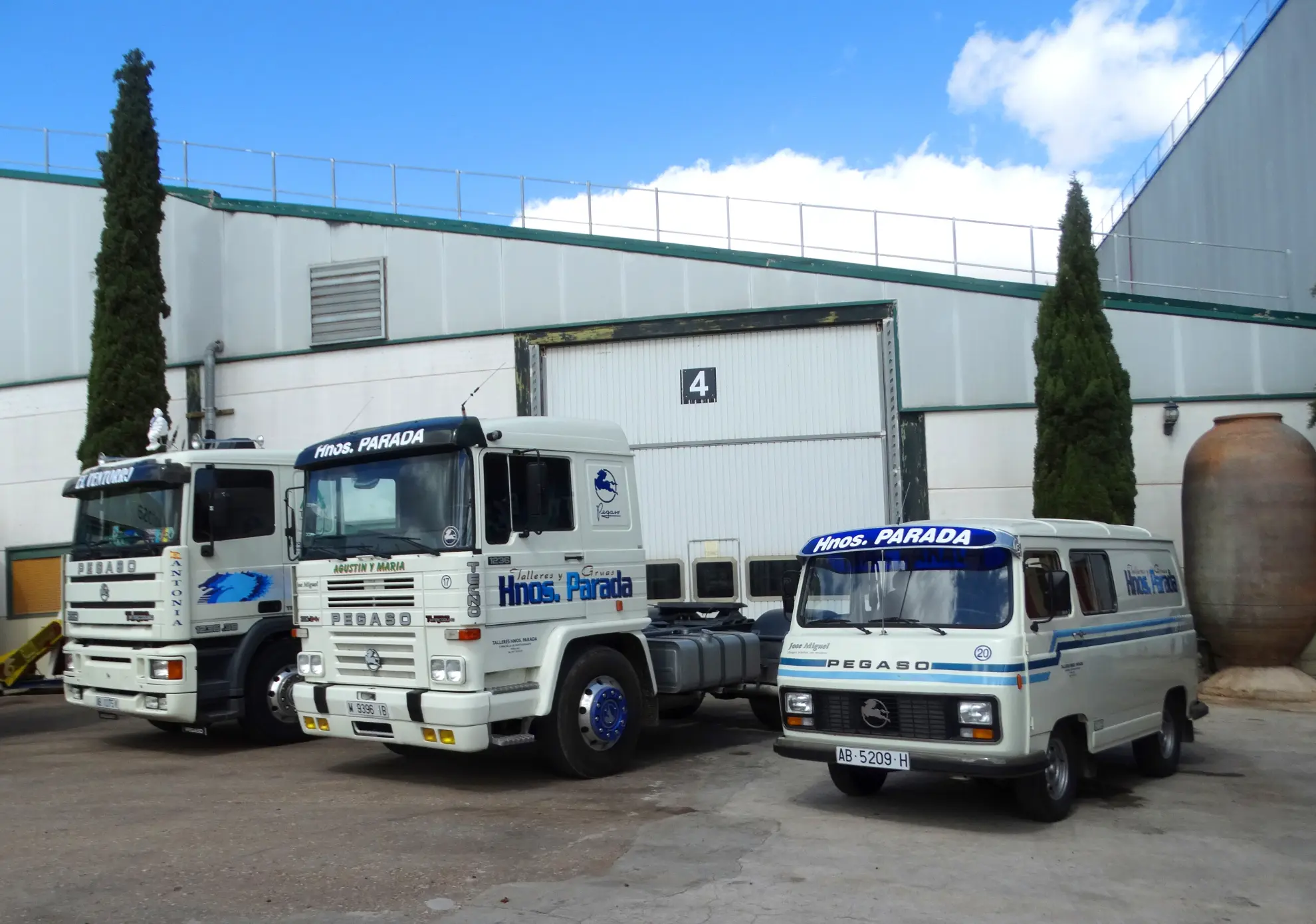 Tres camiones clásicos blancos con la etiqueta "Hnos. Parada" están aparcados frente a un almacén con el número 4 en la puerta, listos para la Vuelta a La Mancha 2025.