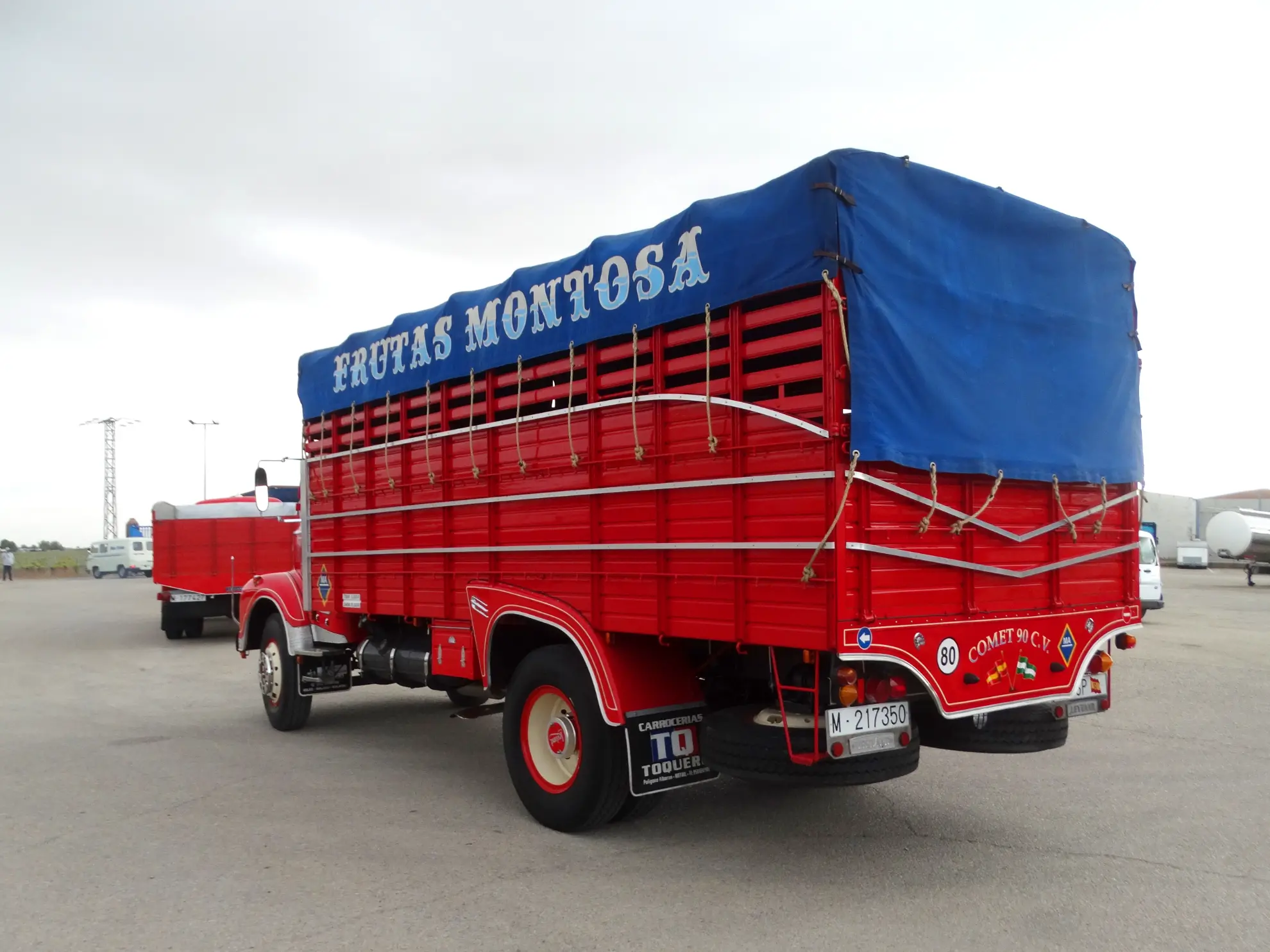 Un camión rojo brillante con una lona azul rotulada "Frutas Montosa" y un cartel visible de "borrador automático" está aparcado en un descampado.