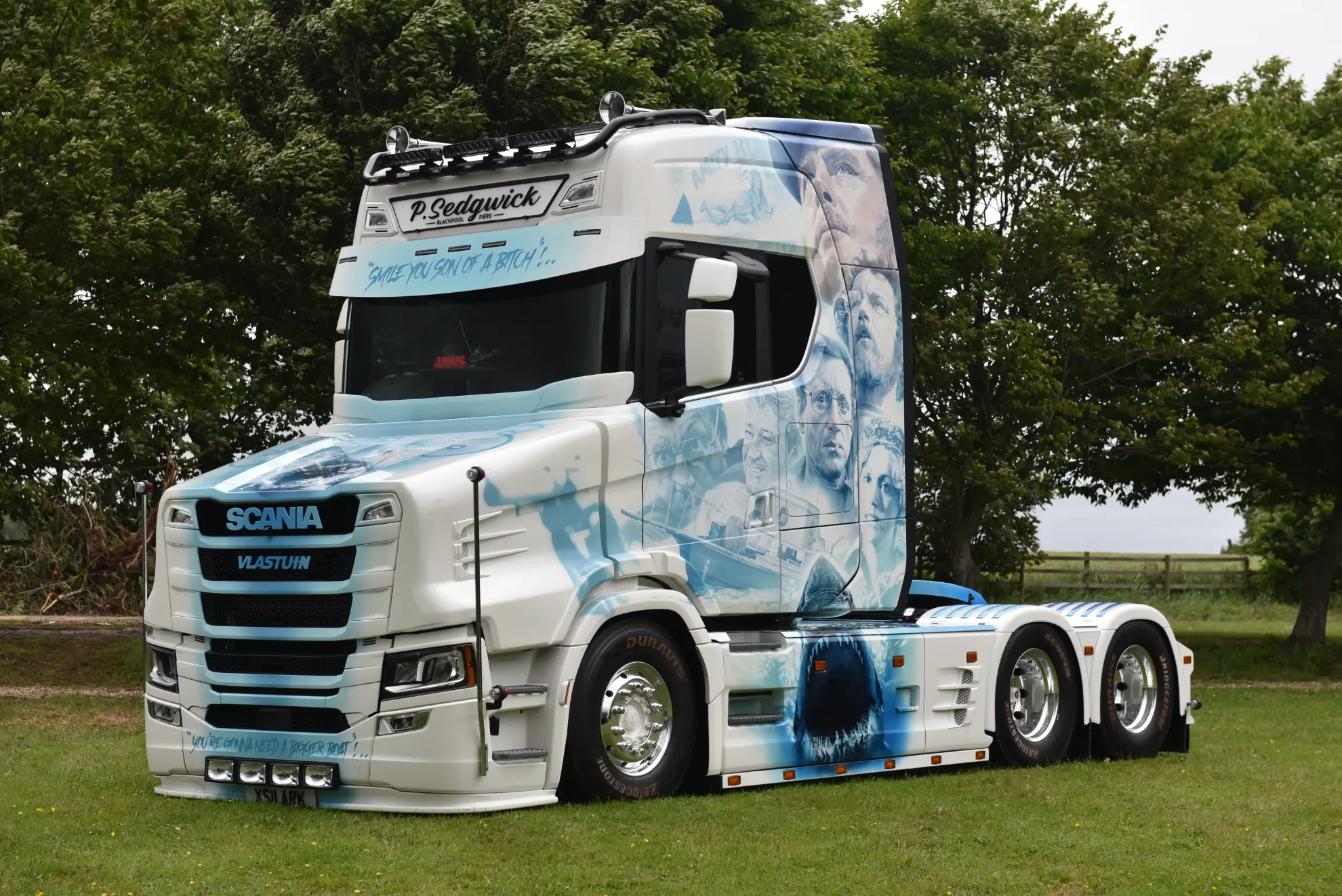 Un Scania 770 S camión blanco customizado con ilustraciones y retratos aerografiados en azul, bautizado como Tiburón, está aparcado sobre hierba cerca de unos árboles.