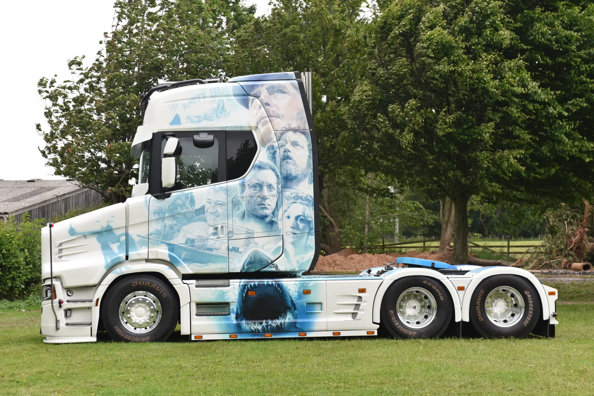 Camión blanco customizado Scania 770 S con retratos en tonos azules pintados en su lateral, aparcado sobre hierba cerca de árboles.