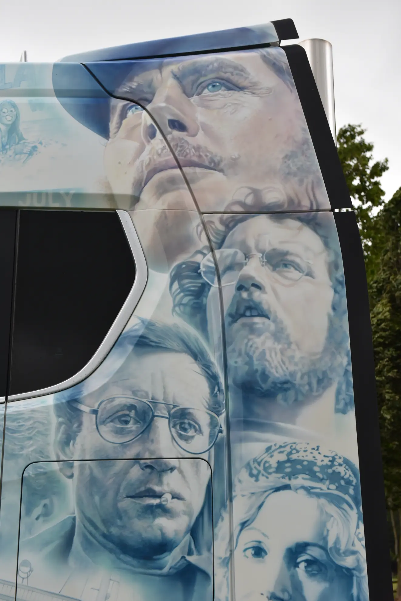 Colorido mural de dramáticos rostros humanos pintado en el lateral de un Scania 770 S, un camión customizado, con árboles de fondo.
