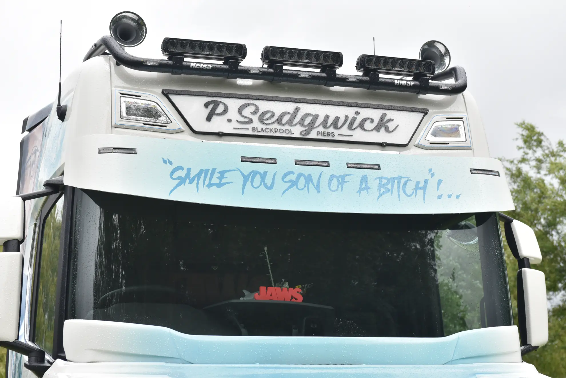Frontal de un camión Scania 770 S blanco con la leyenda "Smile you son of a bitch!" en azul sobre el parabrisas, luciendo este camión customizado inspirado en Tiburón.