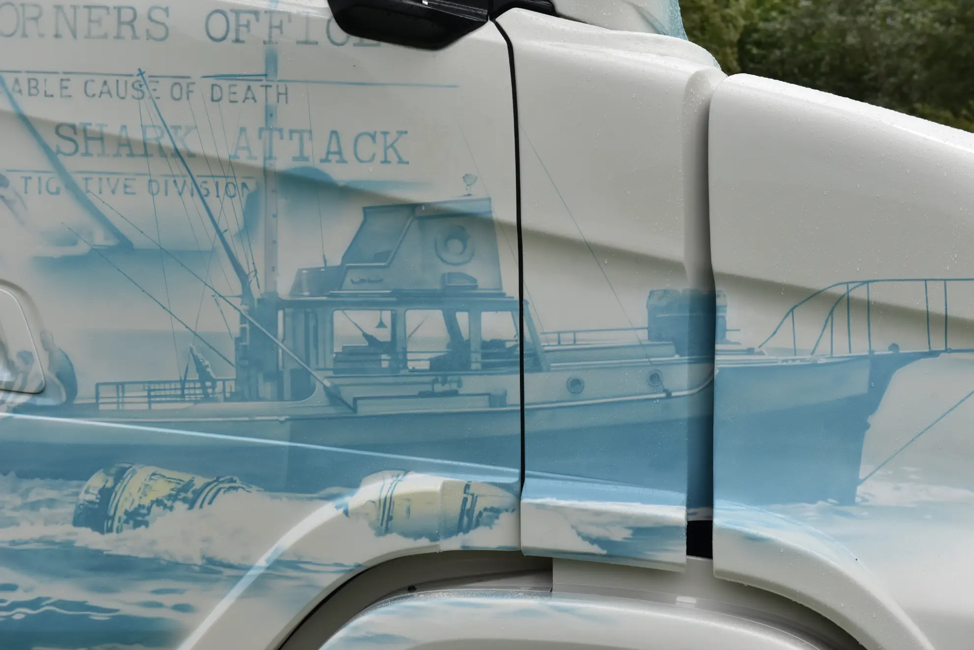 Primer plano de un camión Scania 770 S personalizado con un mural de un barco pesquero en el agua y texto difuminado sobre un ataque al Tiburón.