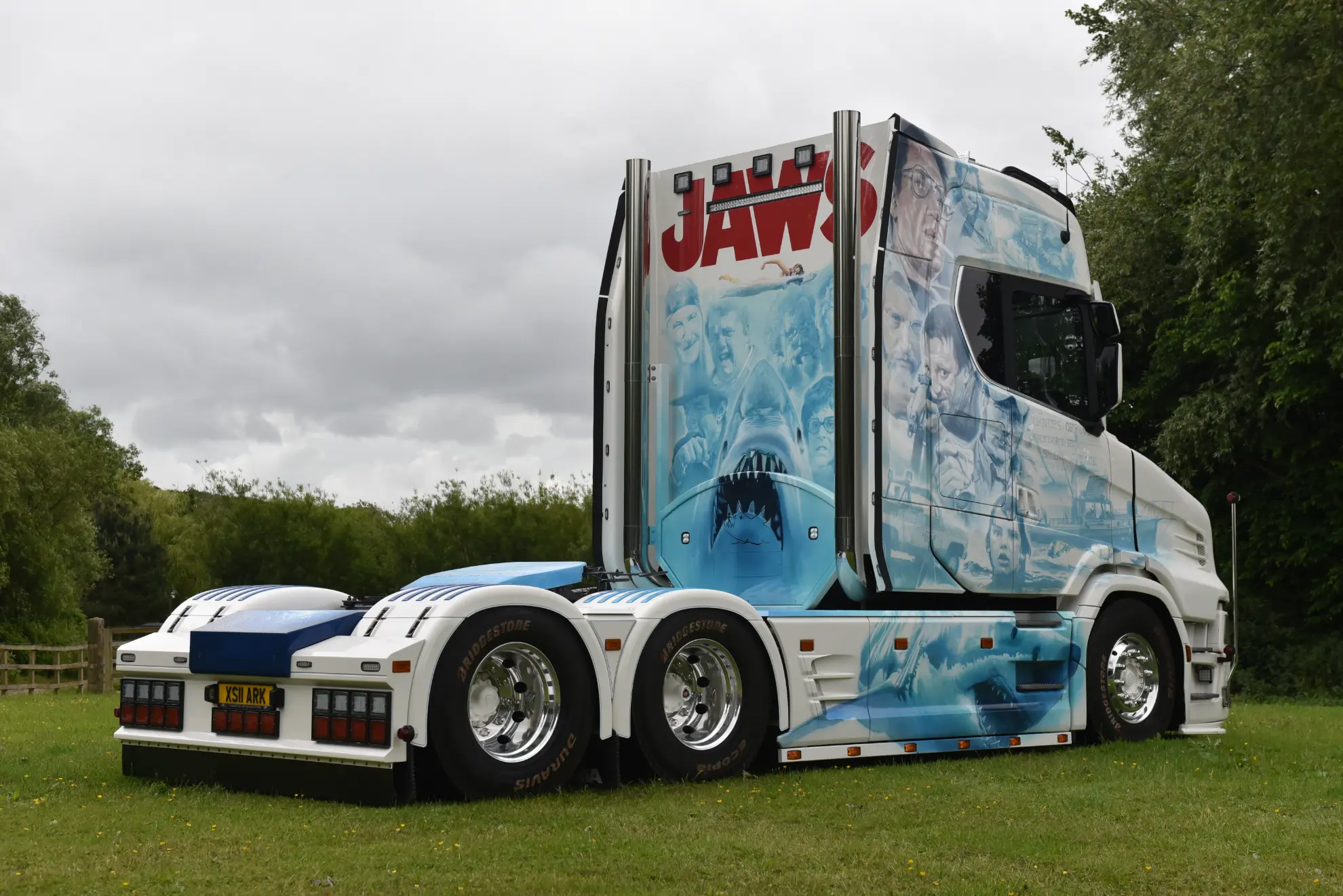 Scania 770 S camión blanco personalizado con un detallado mural del Tiburón "Tiburón", aparcado sobre hierba con árboles al fondo.