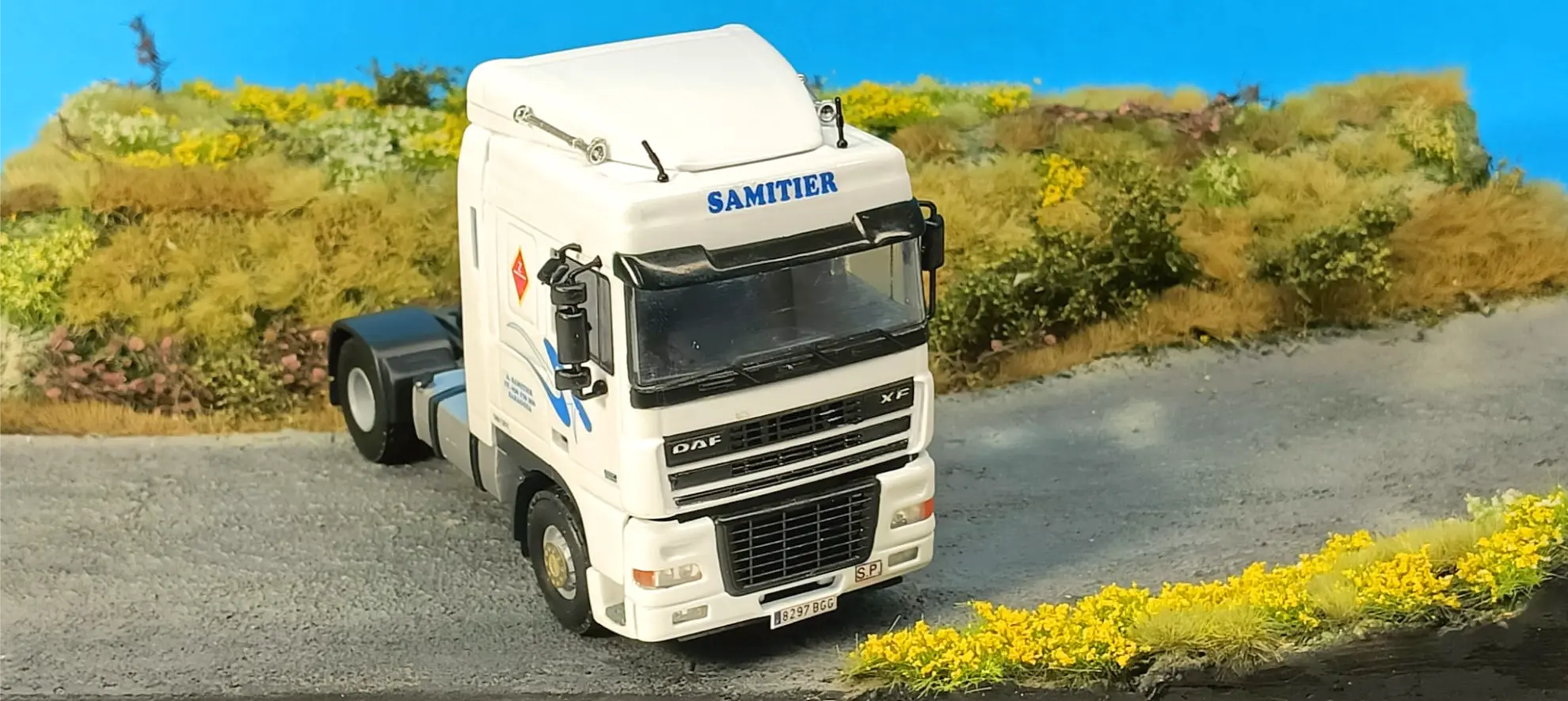 Un camión DAF blanco con la inscripción "SAMITIER" en la cabina está aparcado en una carretera junto a hierba y flores amarillas, lo que lo convierte en la inspiración perfecta para las maquetas de camiones y los interesados en el coleccionismo de camiones.