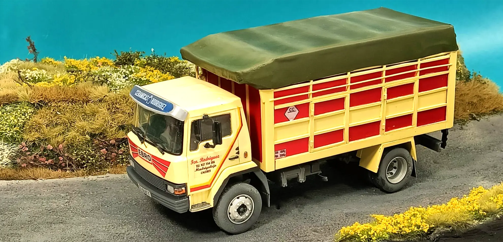 Un camión de carga amarillo y rojo con cubierta verde circula por un camino rural con vegetación alrededor, parecido a maquetas de camiones o a un camión Italeri réplica camión personalizado.